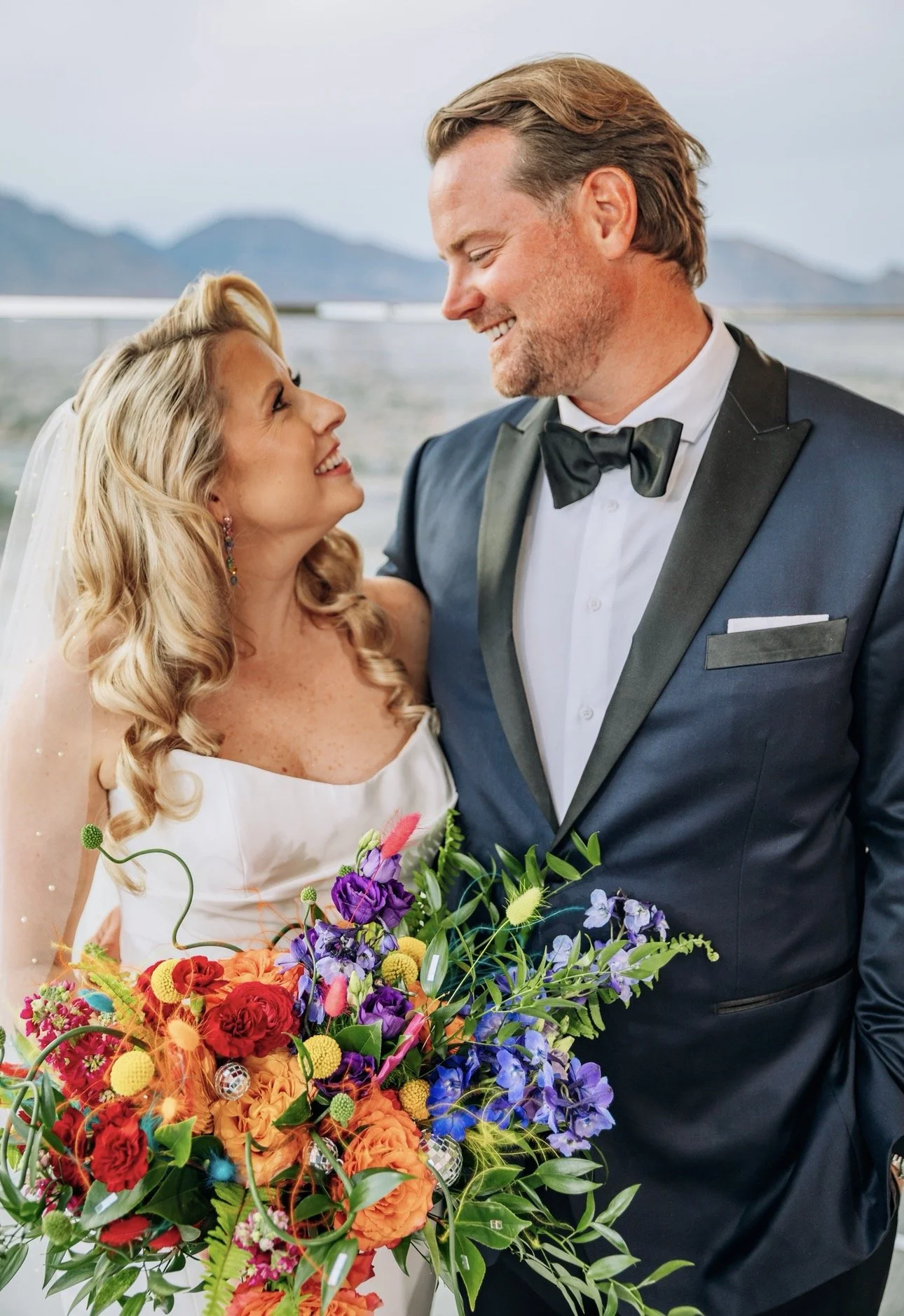 A bride and groom on their wedding day, smiling and looking into each other's eyes, with a colorful bouquet of flowers in front of them.