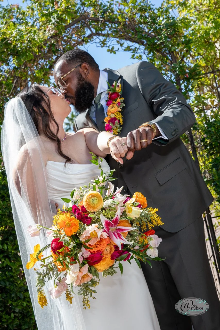 A bride and groom kiss during their outdoor wedding, with the bride holding a large colorful bouquet of flowers and the groom wearing a gray suit with a floral boutonniere.