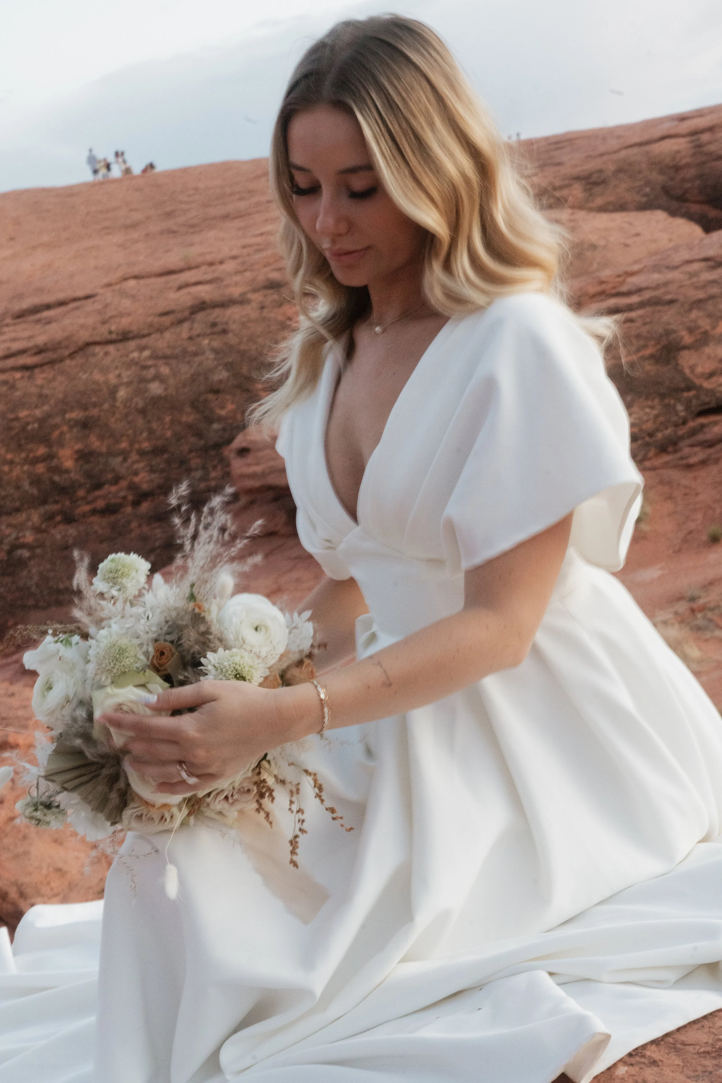 A woman in a white dress sitting on reddish-brown rocks holding a bouquet of white and beige flowers, with a pale sky in the background.