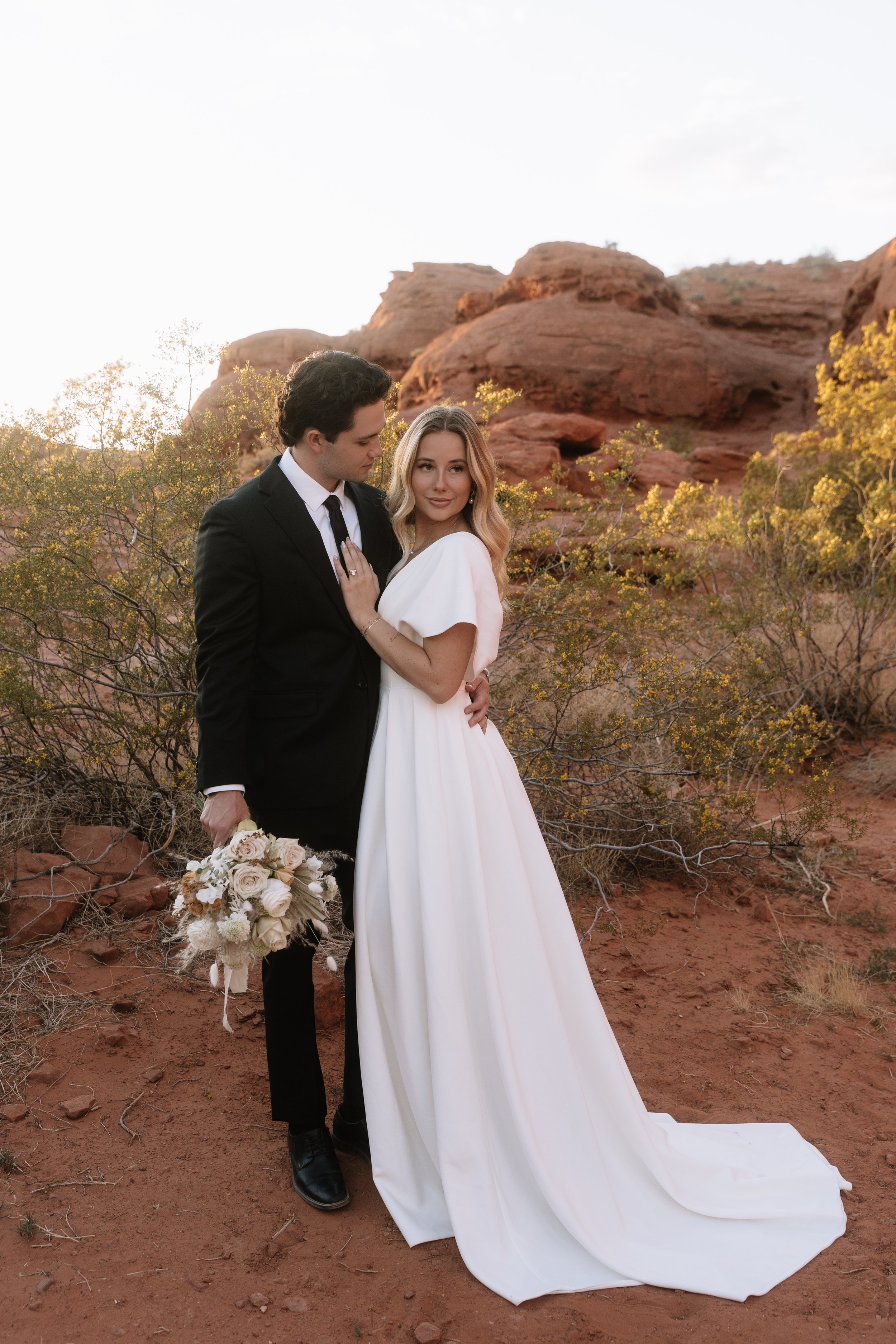 A bride and groom stand together in a desert landscape with red rocks and sparse yellow-green bushes. The groom, dressed in a black suit and tie, holds a bouquet of white roses and other flowers. The bride, wearing a flowing white wedding gown, gazes
