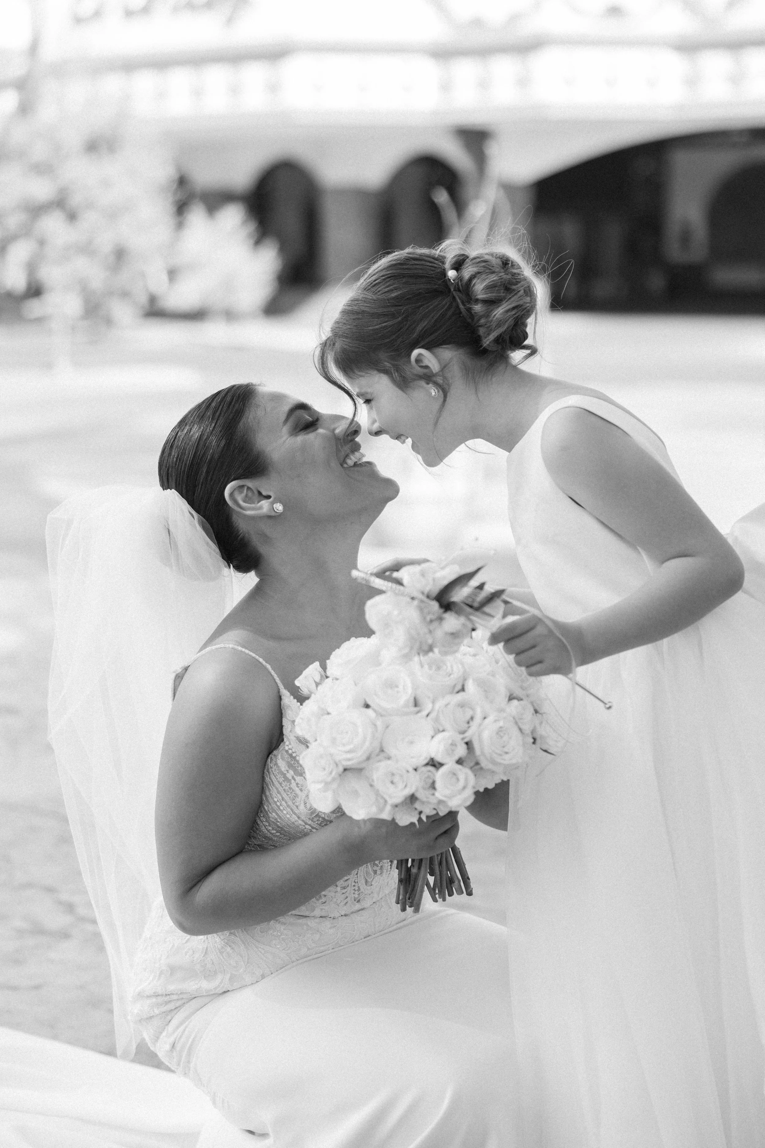Two women, presumably bride and bride's friend, share a joyful moment close to each other outdoors during a wedding, with the bride holding a bouquet of roses.