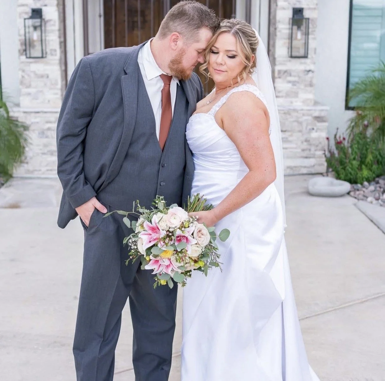 A bride and groom are shown in a close embrace, with their foreheads touching, standing outside a stone building. The bride is holding a bouquet of pink lilies and white roses, and is wearing a white wedding dress. The groom is in a gray suit, white 