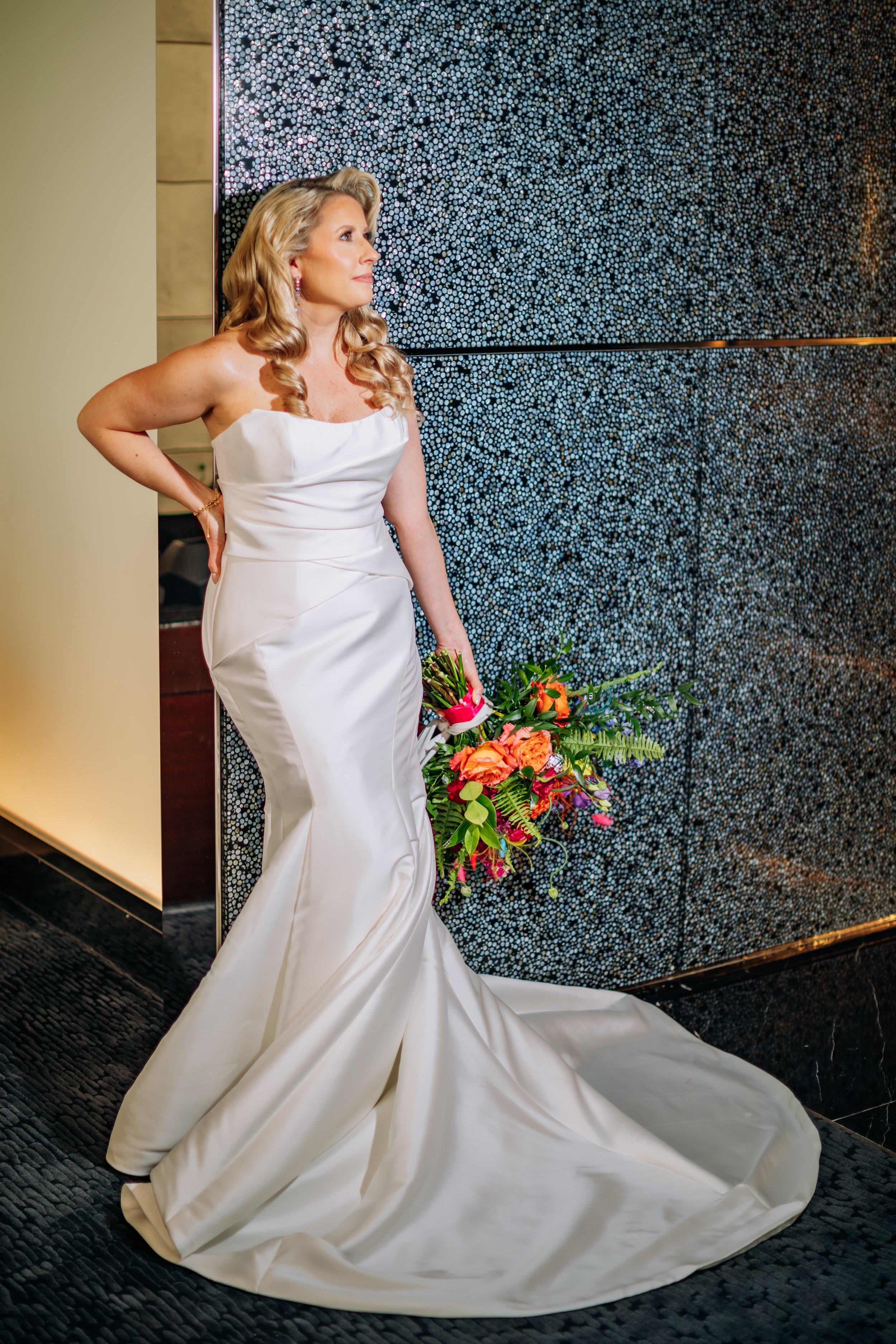 Bridgerton-inspired bride in a white satin wedding gown holding a colorful bouquet, standing against a textured dark wall.