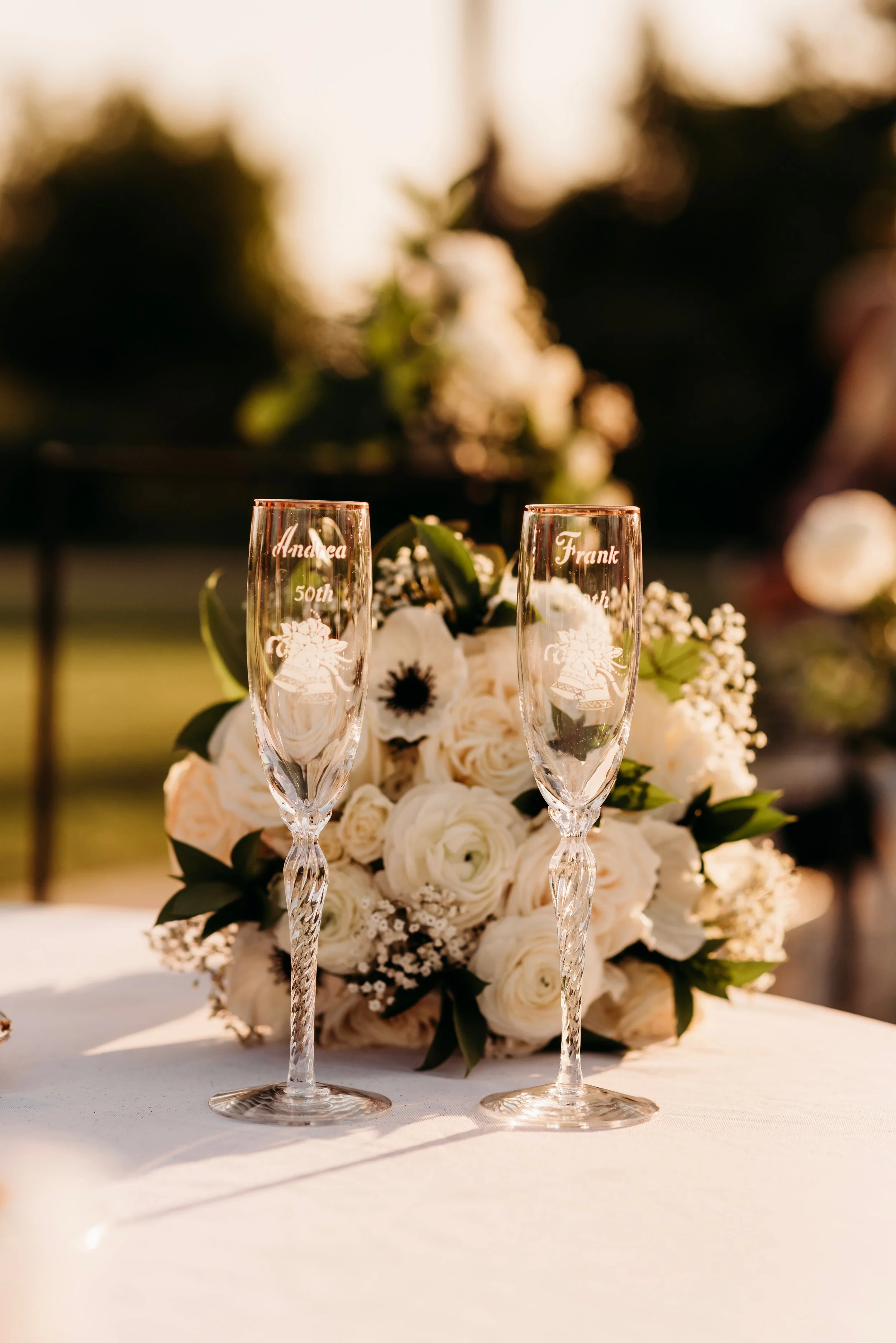 Two engraved champagne flutes with names 'Andrea 50th' and 'Frank 50th' in front of a white flower bouquet on a table at sunset.