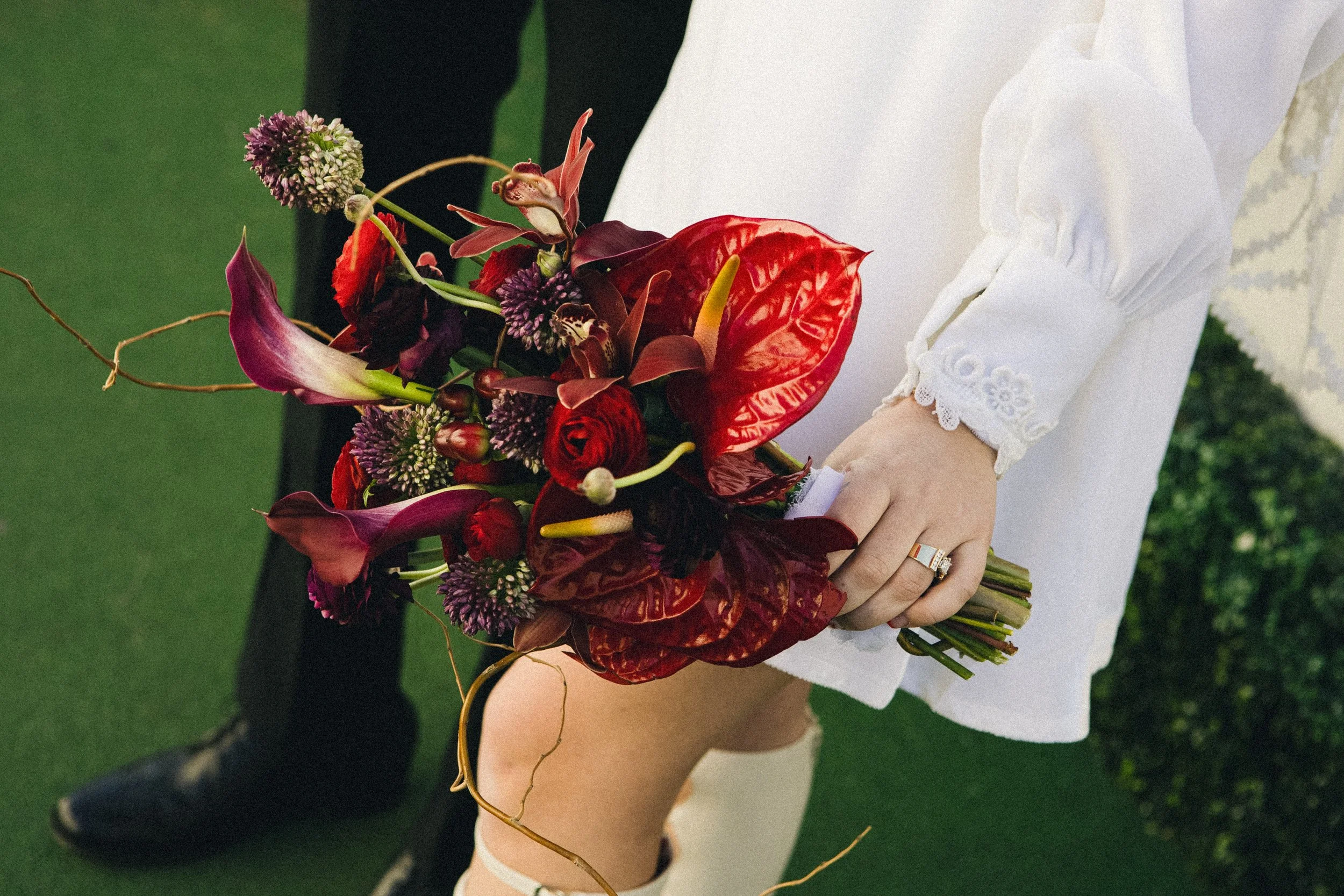 Person holding a bouquet of dark red, purple, and green flowers with a white sleeve and ring on their finger, standing on green grass.
