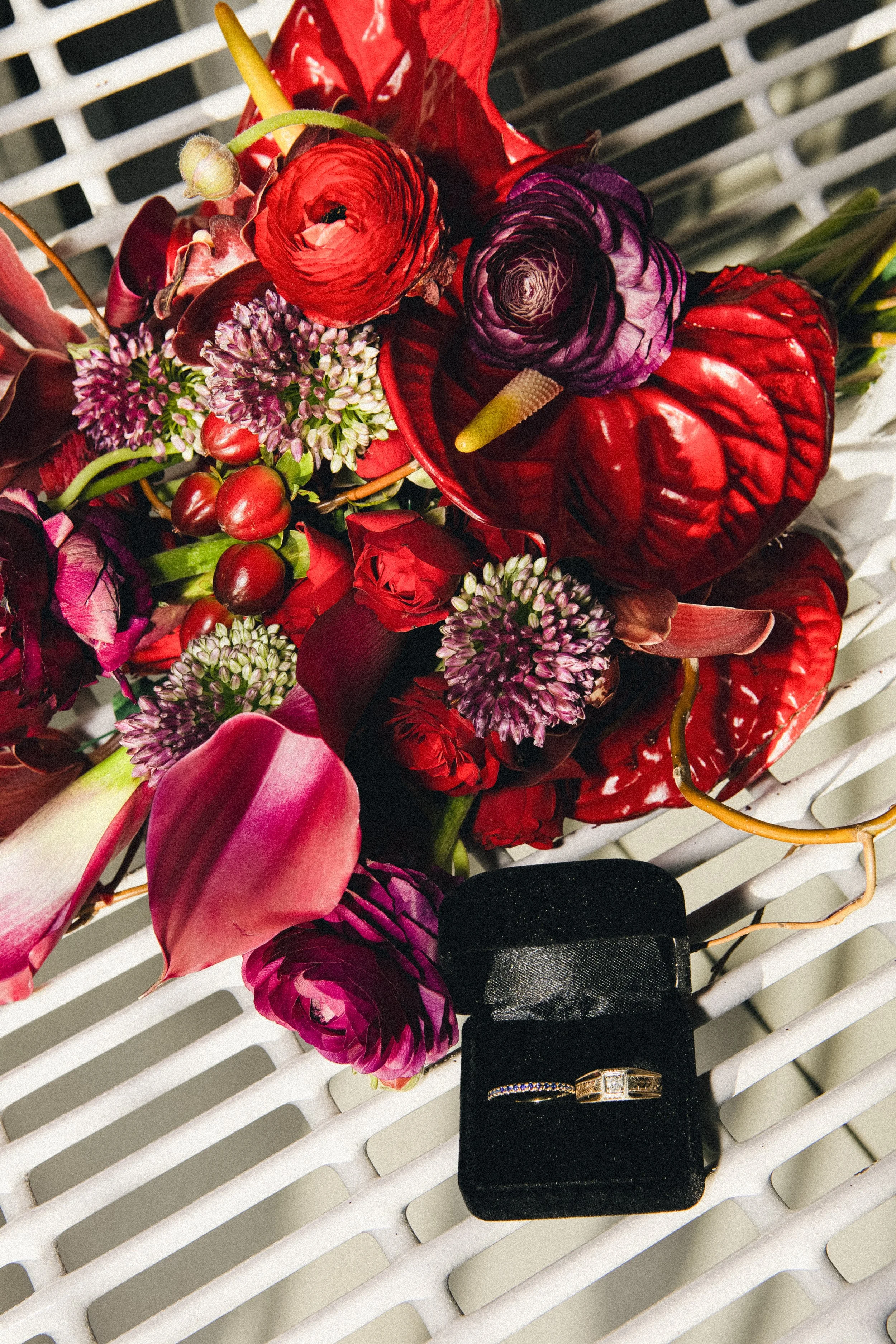 A bouquet of red and purple flowers with green stems, placed on a white lattice surface, alongside two rings in a black ring box, one with a large square diamond and the other with small stones.