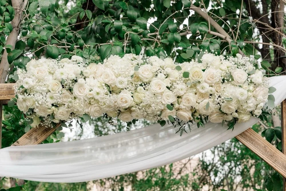 A wedding floral arrangement with white roses, baby's breath, and greenery on a wooden arch draped with white fabric, outdoors among trees.
