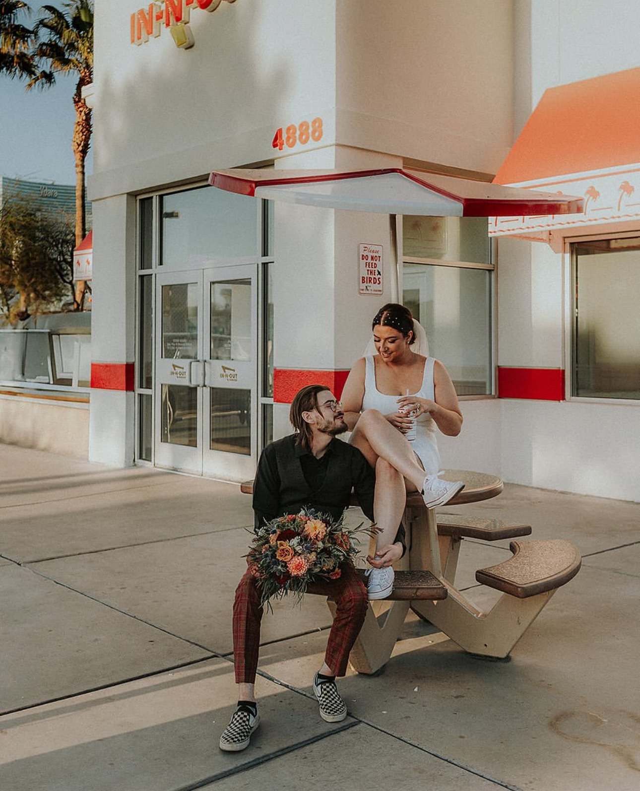 A couple sitting outside a fast-food restaurant, with one person sitting on a picnic table and the other sitting on their lap. The person on the lap is holding a drink, and the person sitting on the table is holding a bouquet of flowers.