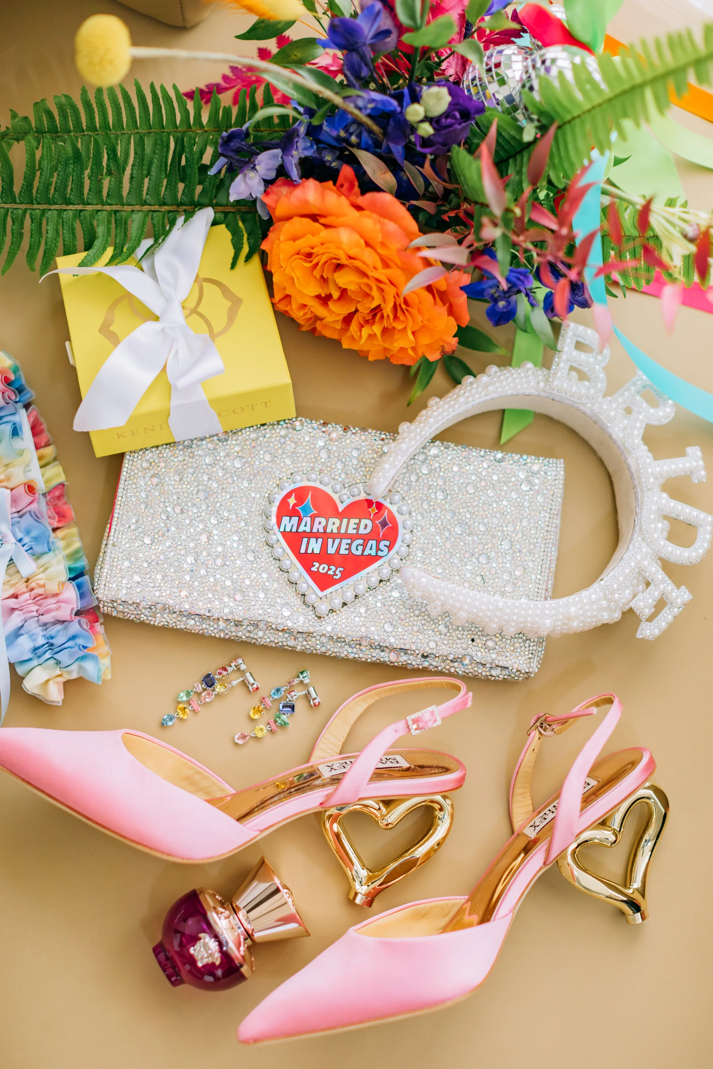A celebratory arrangement including a large bouquet of colorful flowers, a yellow gift box with a white ribbon, a glittery clutch bag with a 'Married in Vegas 2025' sticker, a white 'Bride' headband, pink high-heeled shoes with gold heart-shaped acce