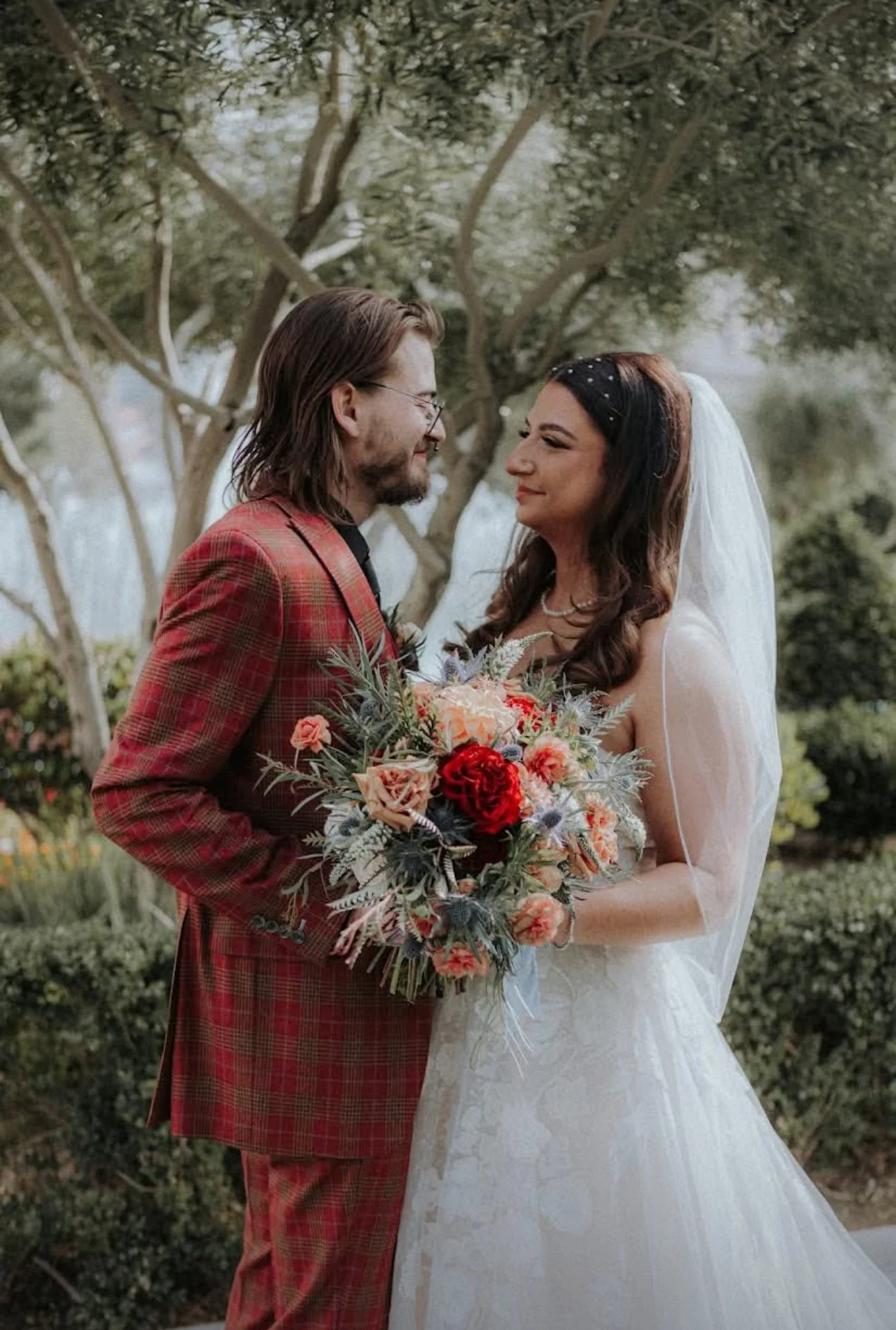 A bride and groom stand close together outdoors, smiling at each other. The groom is wearing a red plaid suit, and the bride is dressed in a white wedding gown with a veil, holding a bouquet of red, pink, and peach flowers with greenery.