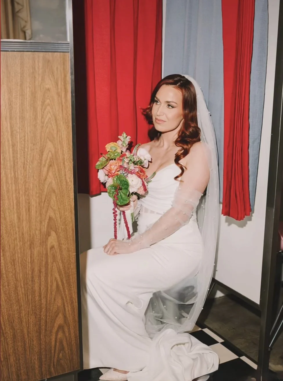 A woman dressed in a white wedding gown sitting in a changing room holding a bouquet of flowers, with red and blue curtains in the background.