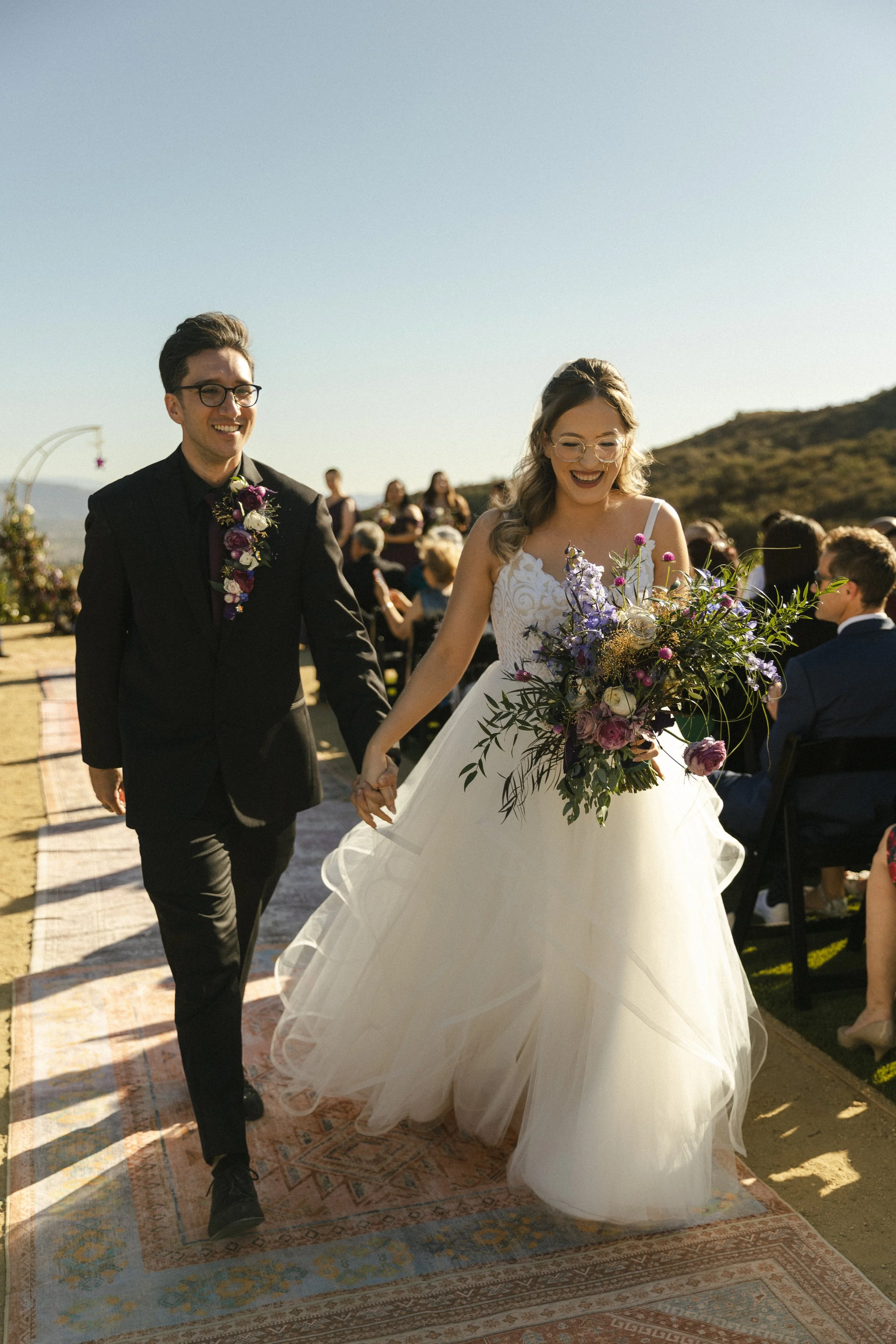 A bride and groom walking hand in hand at their outdoor wedding ceremony, with guests seated in the background and a scenic hillside under a clear sky.