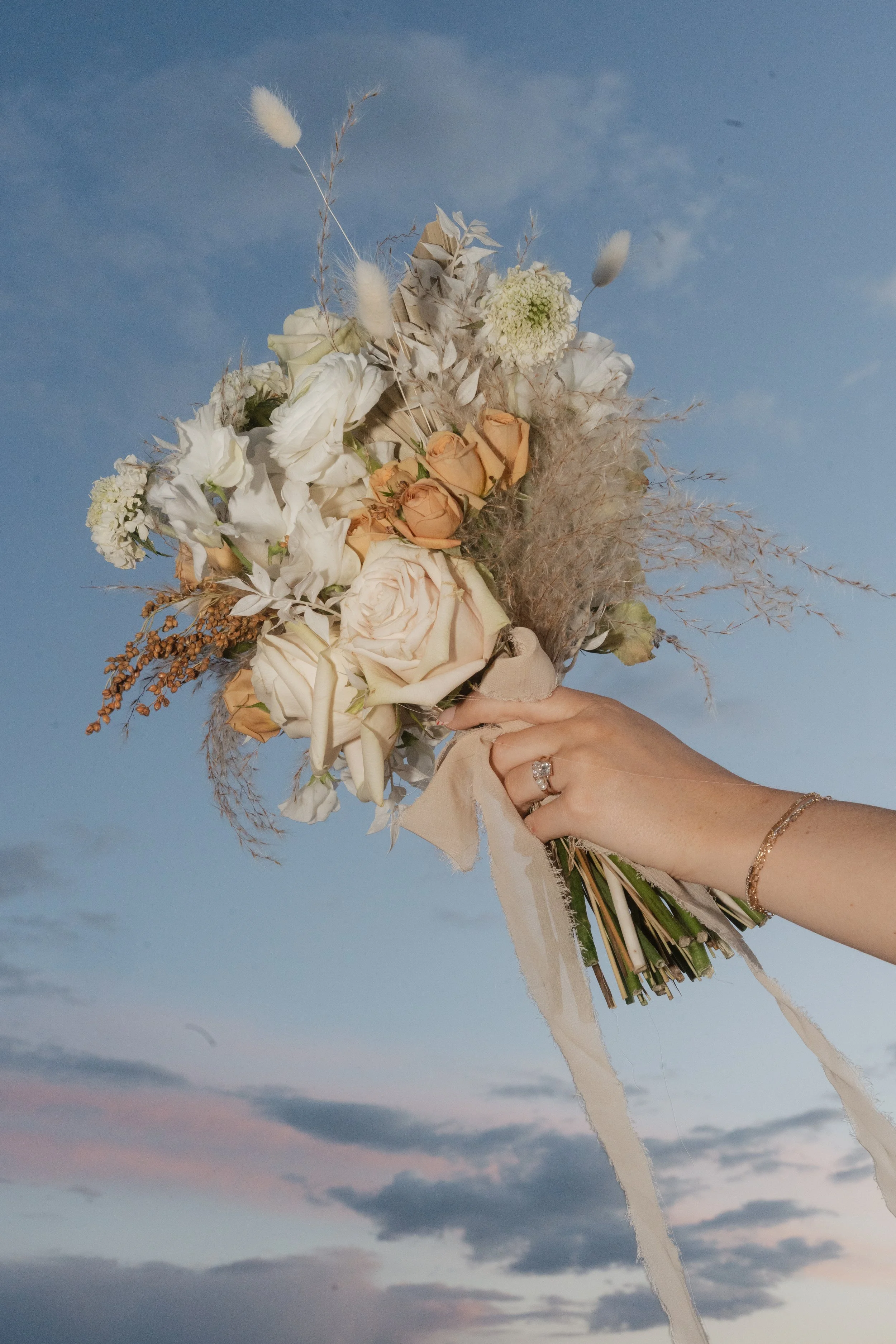 A person's hand holding a bouquet of white and beige flowers against a sky with clouds at sunset.