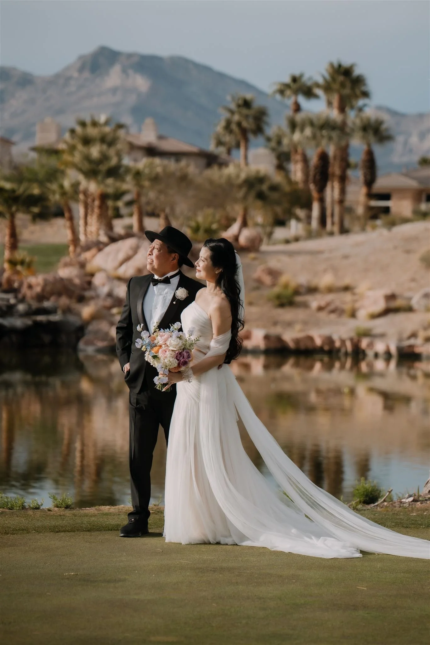 A bride and groom standing by a pond with mountains and palm trees in the background. The bride is wearing a white wedding gown with a long train and holding a bouquet, while the groom is dressed in a tuxedo with a flower boutonniere and a wide-brimm