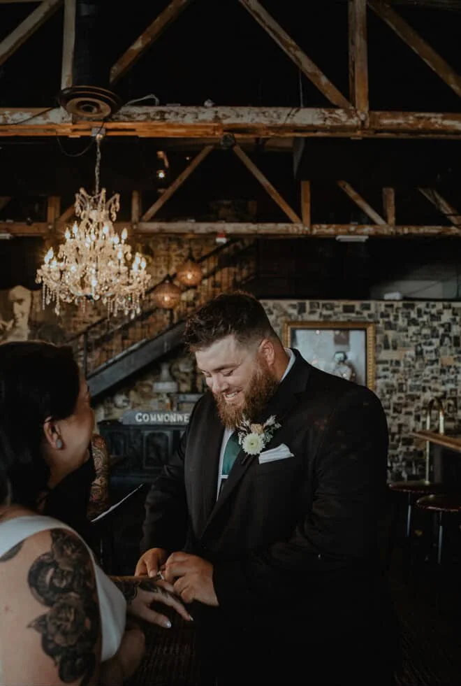 Man in a black suit and tie exchanging rings with a woman in a white dress during a wedding ceremony inside a rustic venue with exposed wooden beams, a chandelier, and a brick wall.