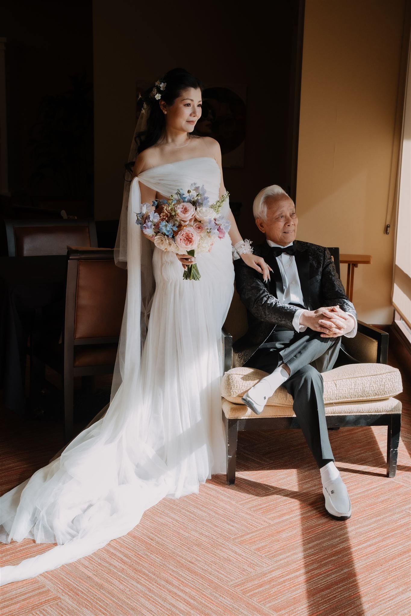 A bride in a white wedding dress holding a bouquet of pink and purple flowers, standing beside a seated elderly man in a tuxedo, possibly her grandfather, in a room with sunlight streaming in.