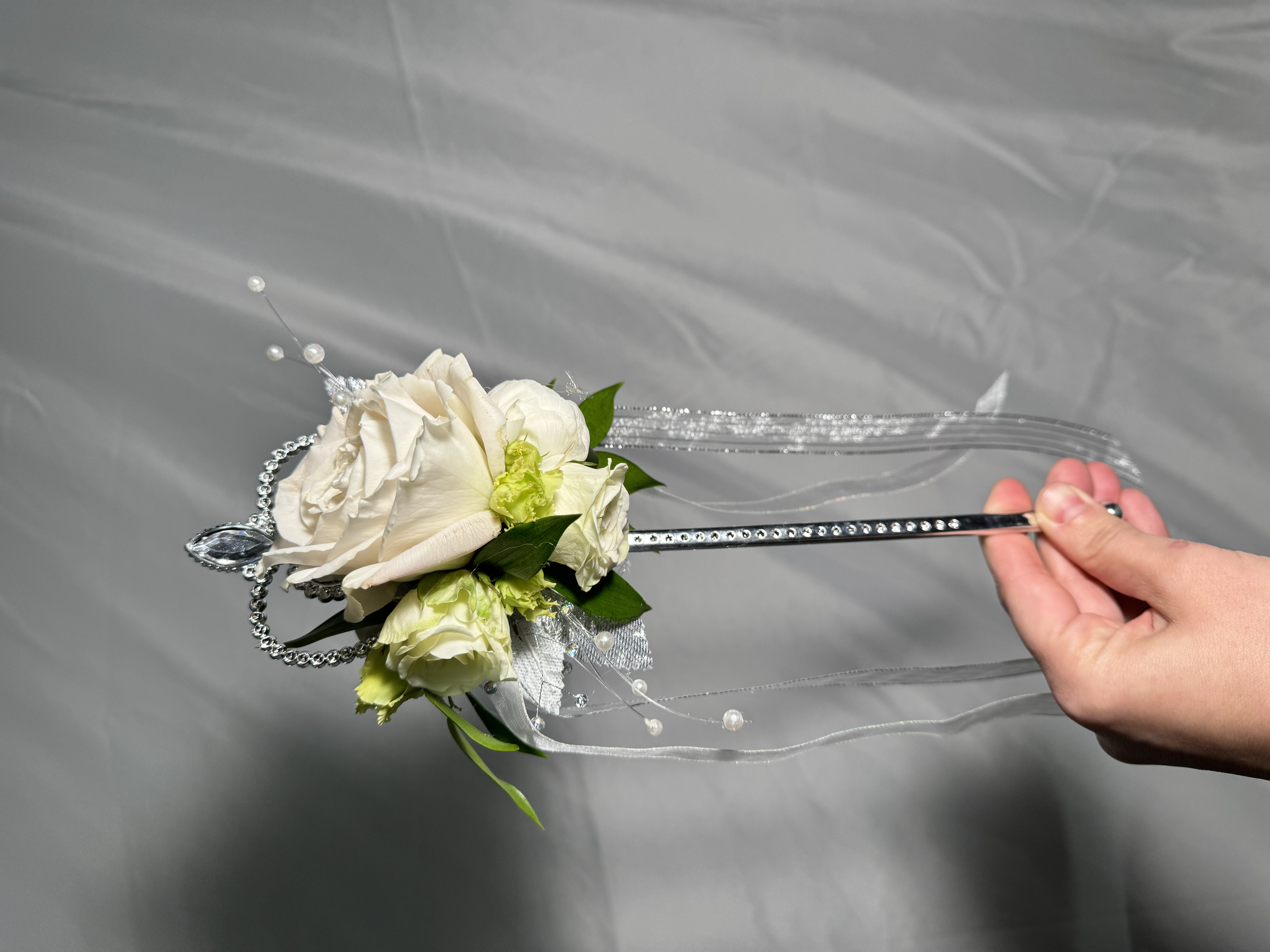 Hand holding a decorative bouquet of white roses, green leaves, and silver embellishments on a gray surface.