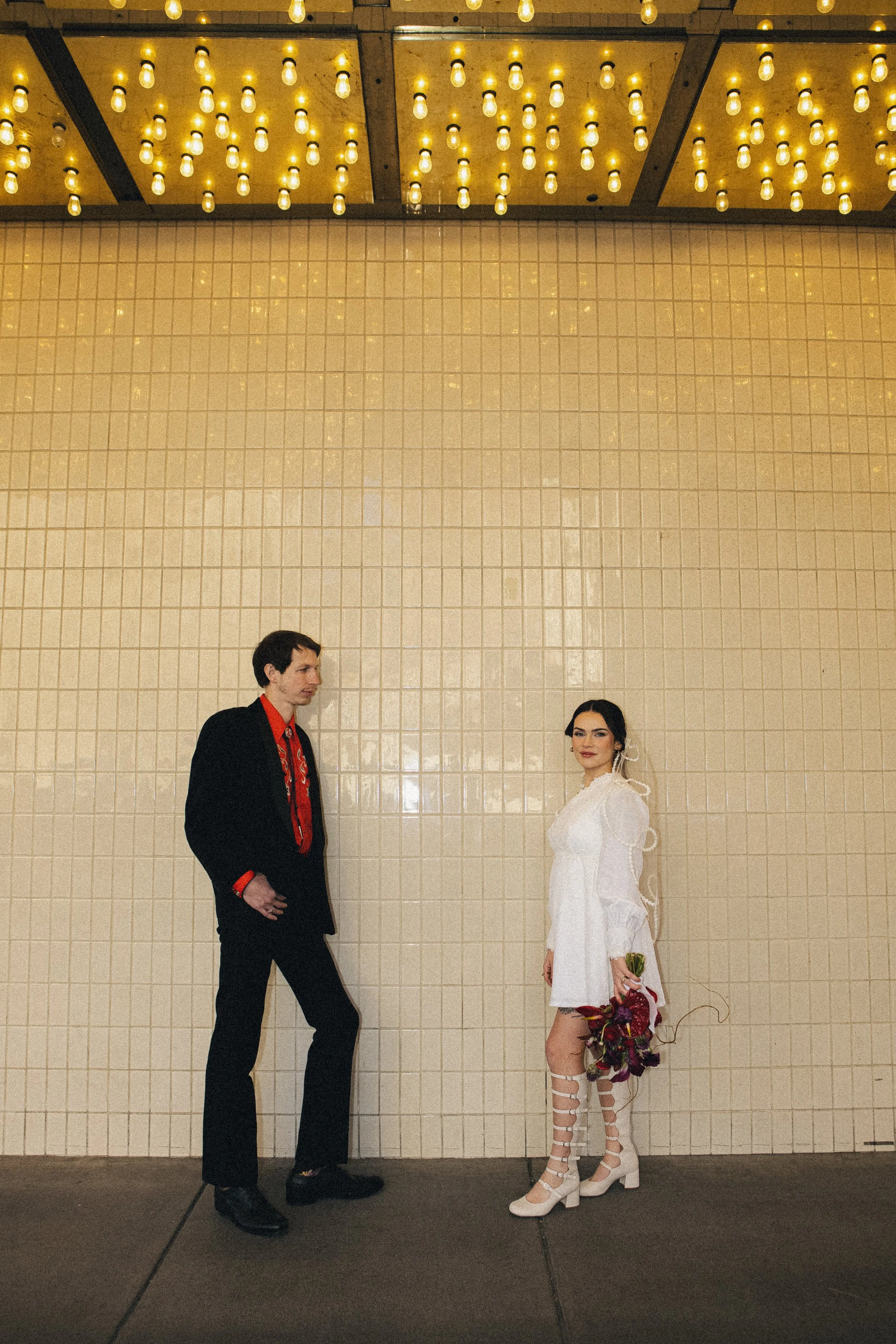 A man and woman dressed in formal attire standing in front of a beige tiled wall with yellow string lights hanging from the ceiling.