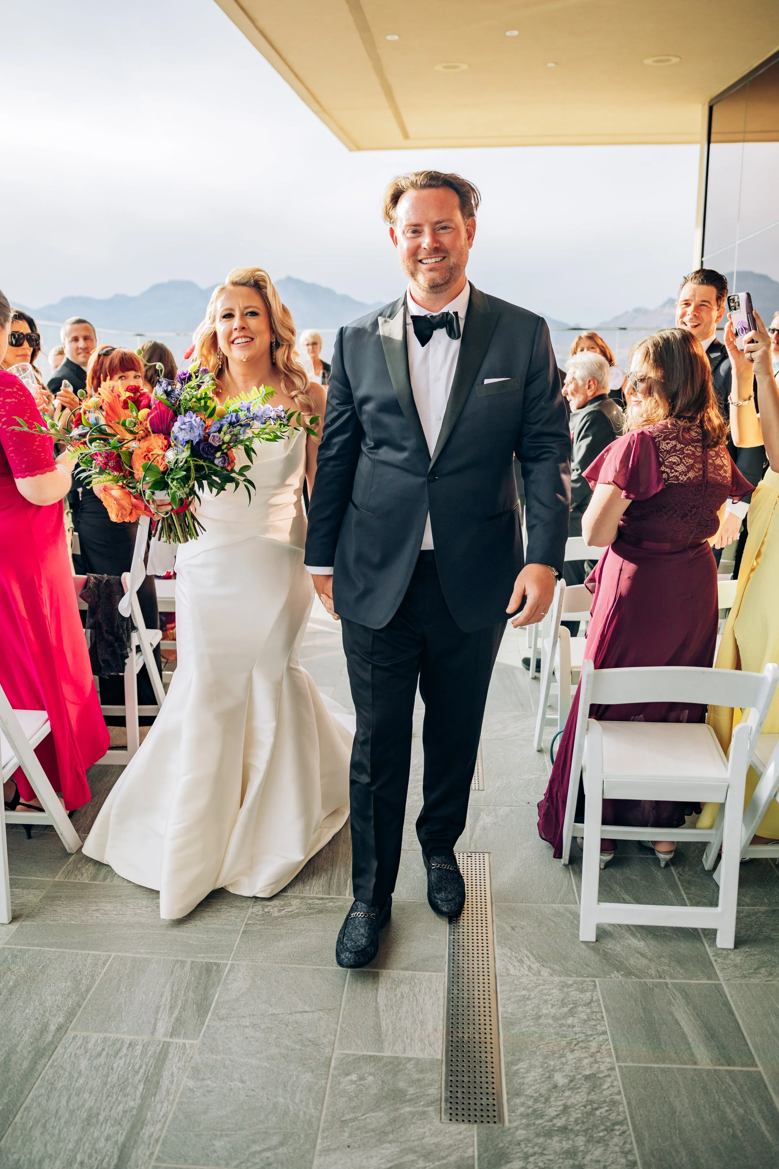 A bride in a white wedding gown holding a bouquet and a groom in a dark suit with a bow tie walking together at an outdoor wedding reception, with guests seated at tables and mountains in the background.