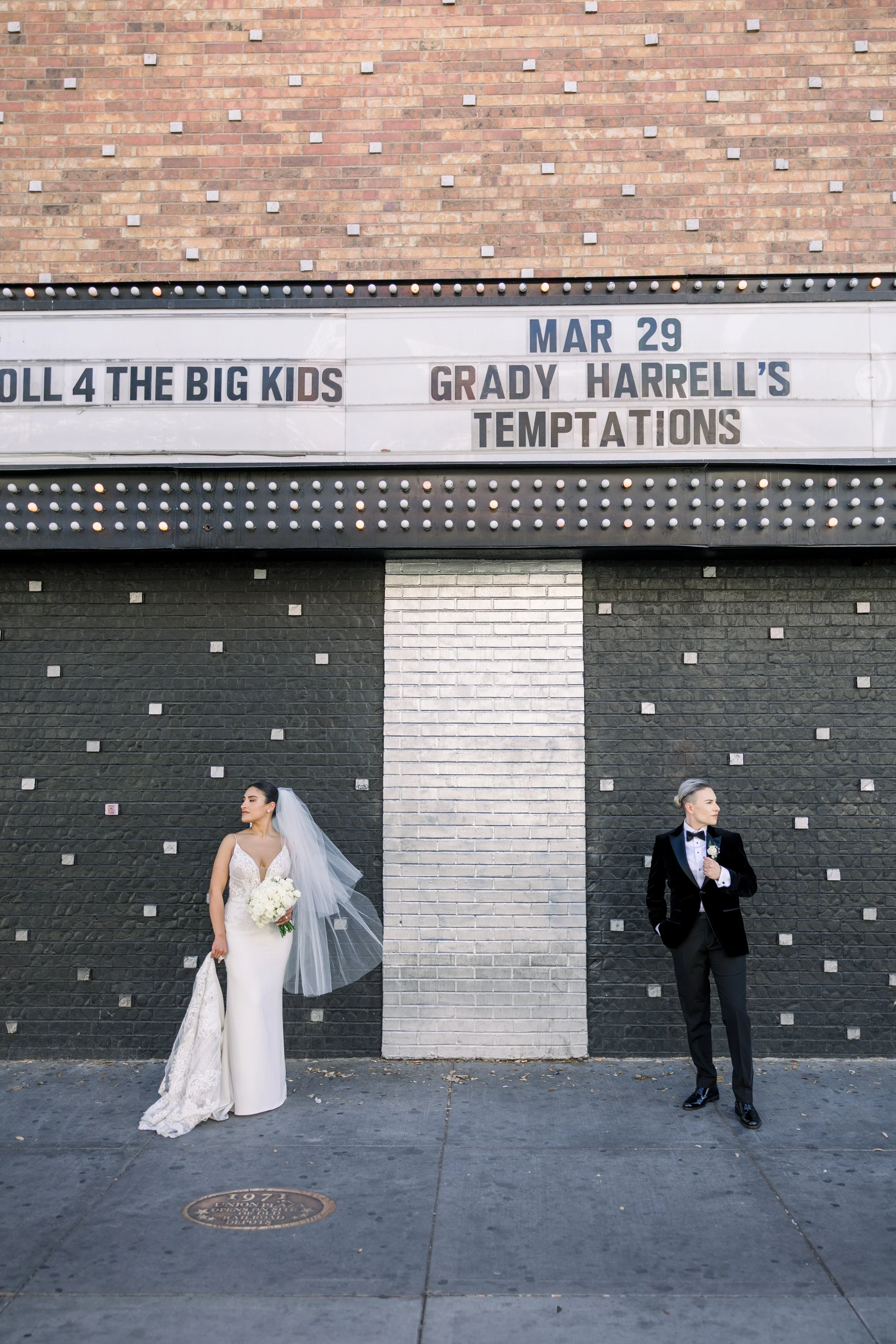 A bride and groom in wedding attire stand on opposite sides of a black and white brick wall in front of a marquee sign that says "March 29, Roll 4 the Big Kid, Grady Harrell's Temptations." The bride is holding a bouquet of white flowers and wears a 