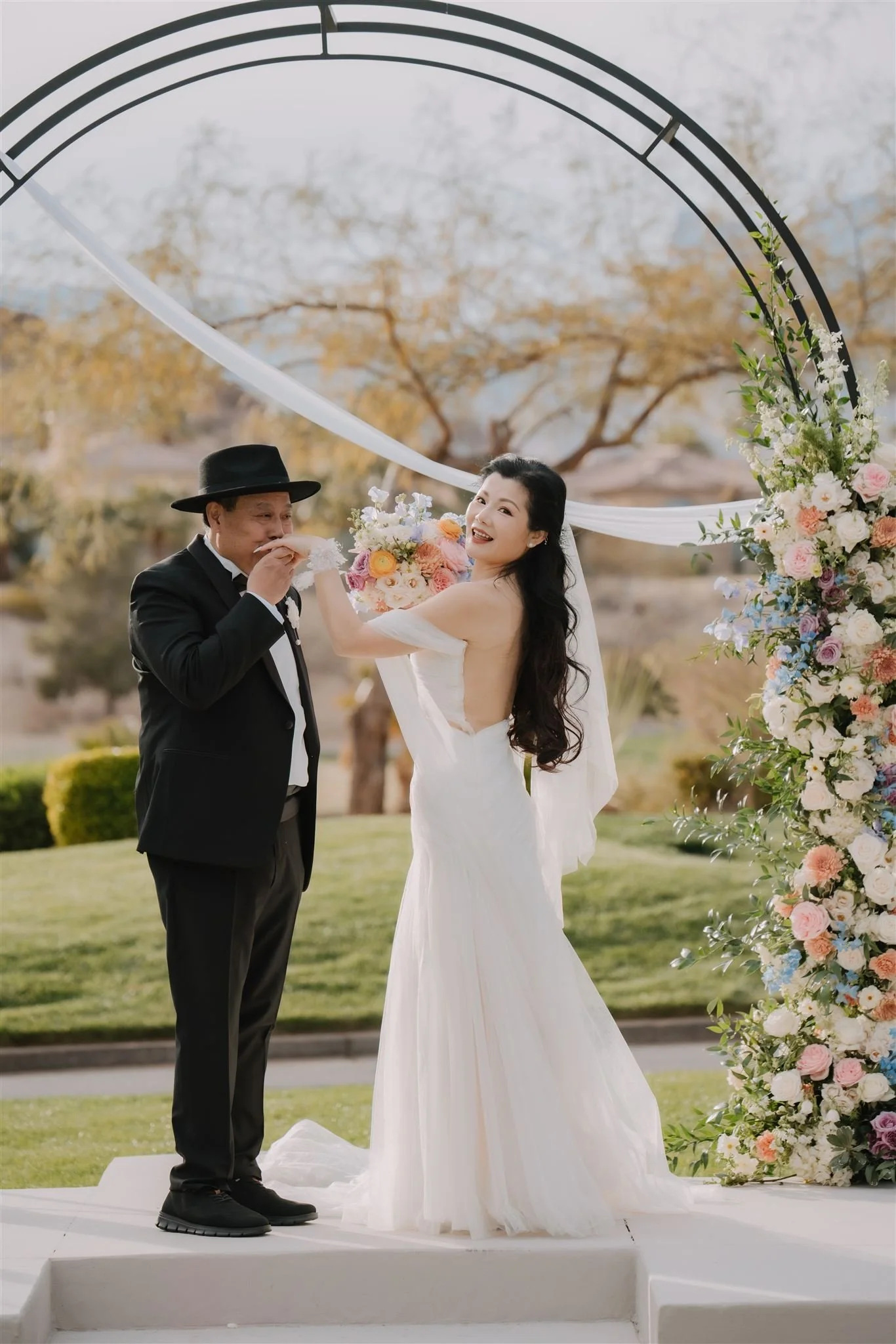 A bride and an groom share a moment during a wedding ceremony outdoors, with an arch decorated with flowers behind them.