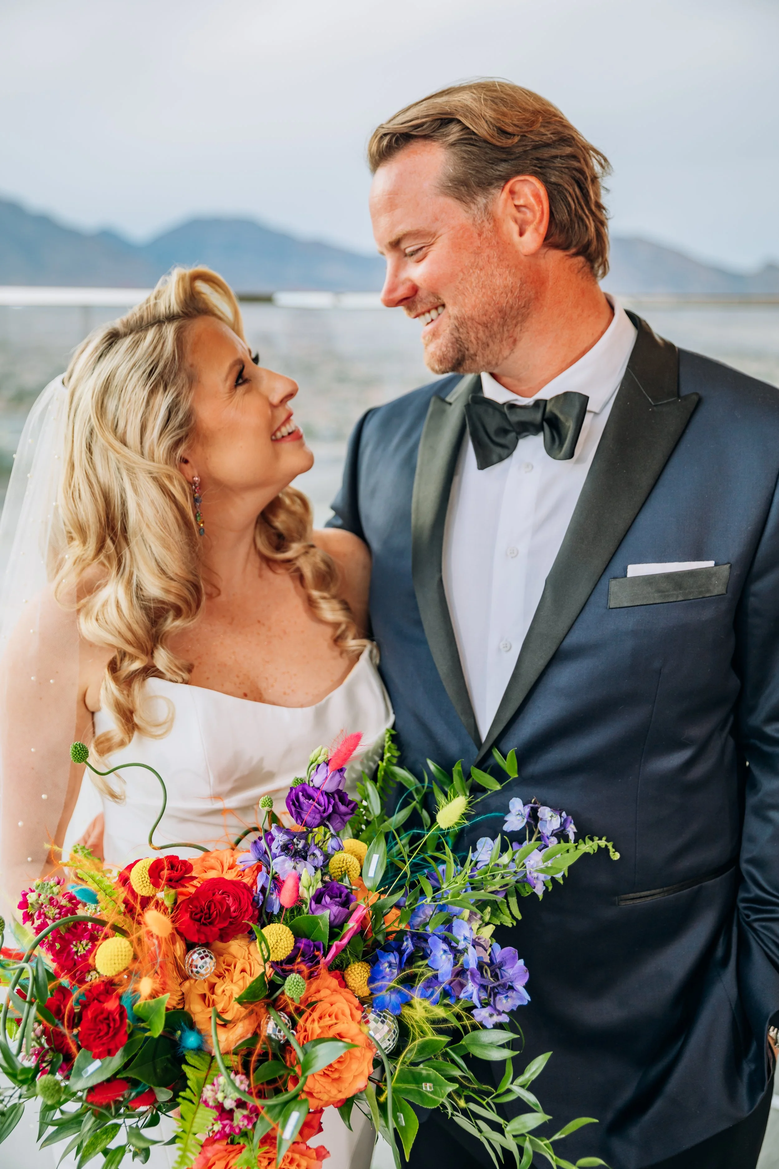 A bride and groom smiling at each other, the bride holding a colorful bouquet of flowers, during their wedding celebration.