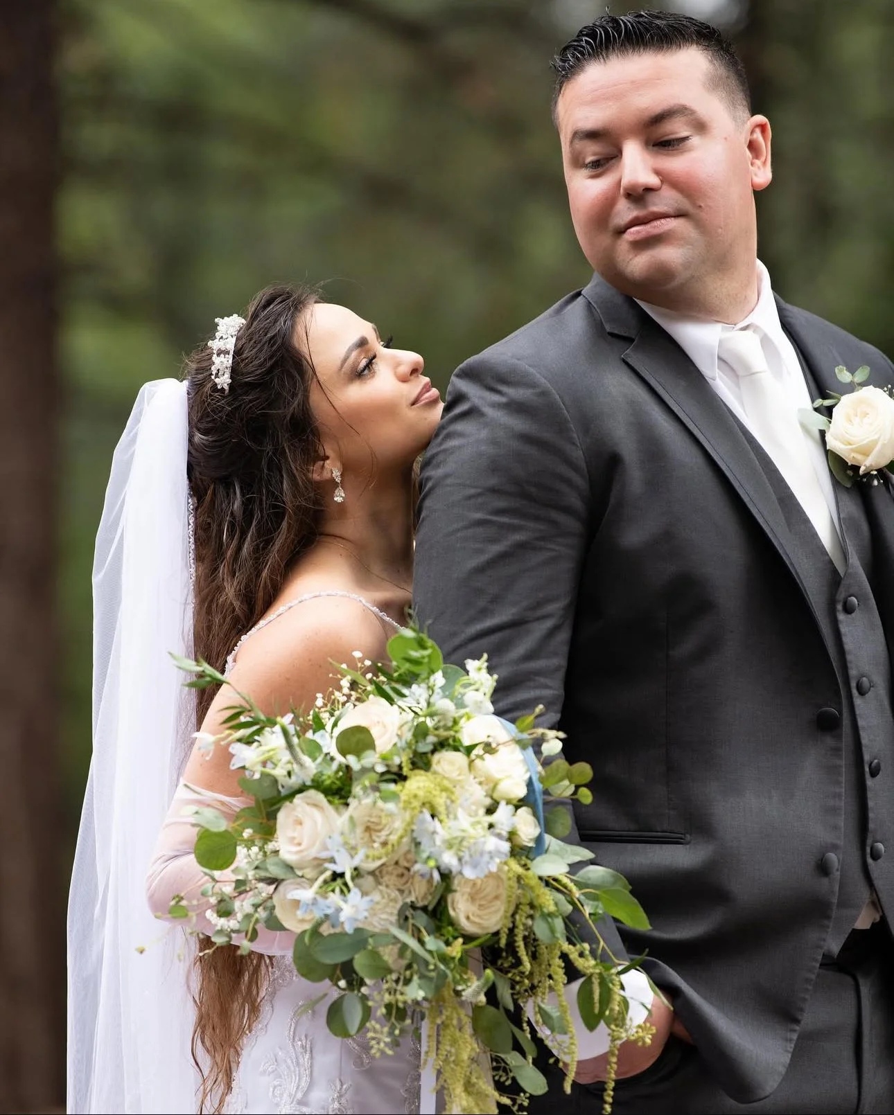 A bride with long dark hair and a tiara stands behind a groom in a gray suit outdoors, holding a bouquet of white and green flowers, as she gazes at him with an affectionate expression.