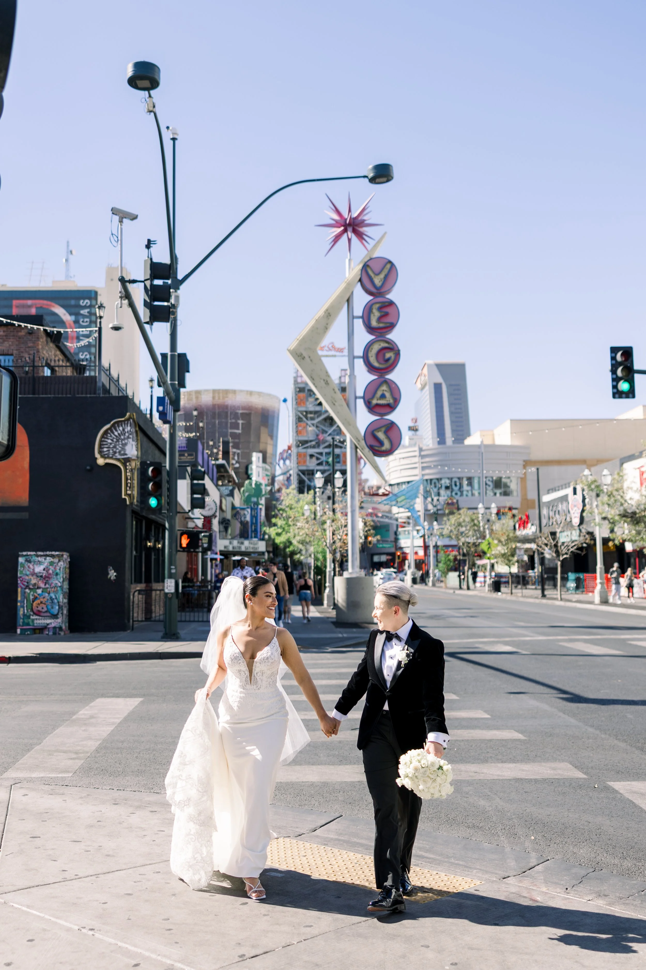 Two brides in wedding dresses holding hands and walking across a city street, with a neon 'VEMOS' sign in the background.