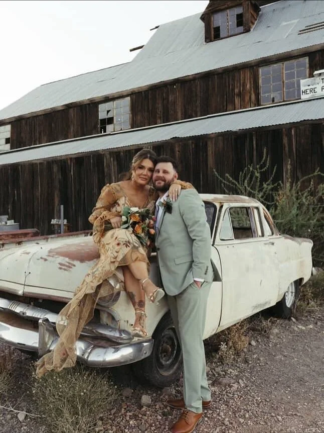 A couple dressed in wedding attire poses in front of an old, rusty car outside a rustic wooden building.