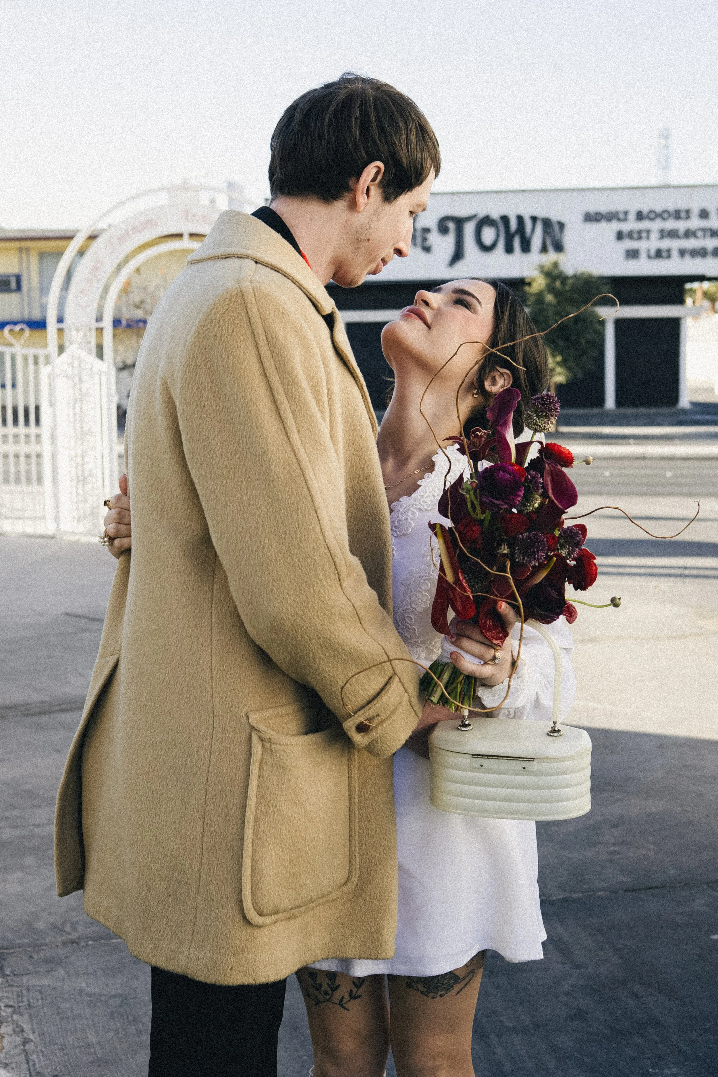 A couple standing closely together on a city sidewalk, looking into each other's eyes. The woman is holding a bouquet of dark red and purple flowers and a white handbag. The man is wearing a beige coat. They appear to be in a romantic moment.