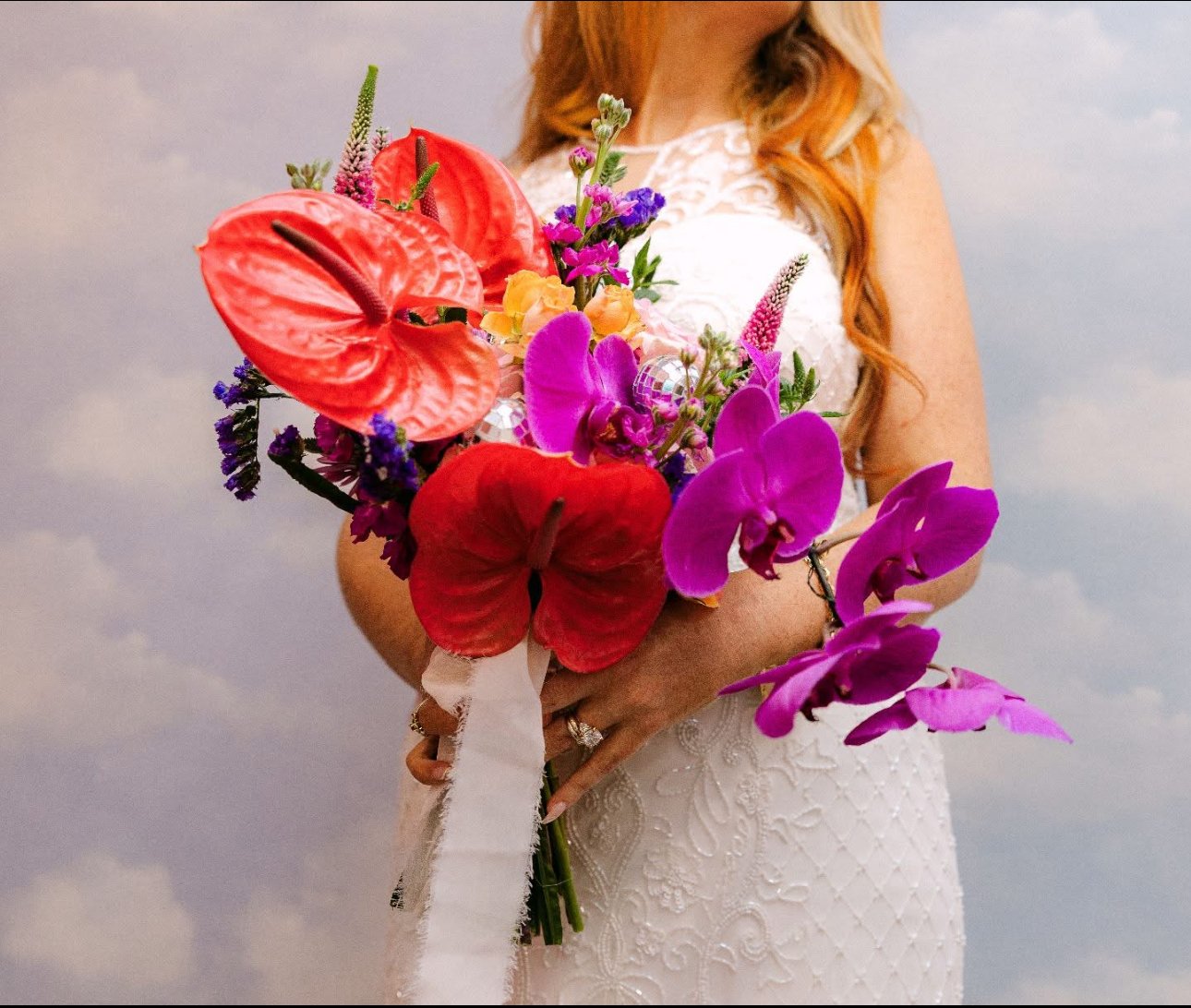 A woman in a white lace dress holds a vibrant bouquet of pink, purple, red, and orange flowers against a cloudy sky background.