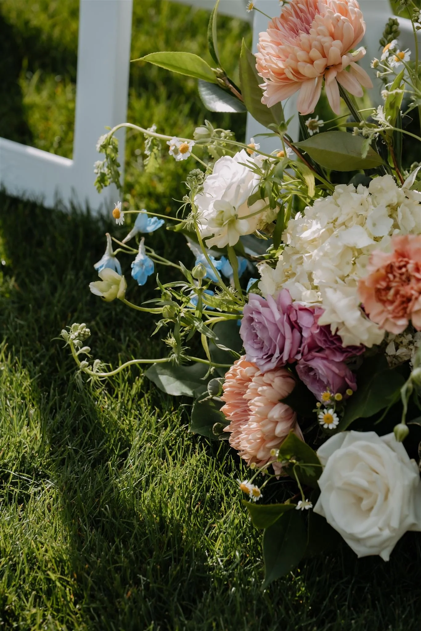 A colorful bouquet of various flowers, including white roses, pink peonies, purple roses, white hydrangeas, and small daisies, lies on green grass near a white chair.