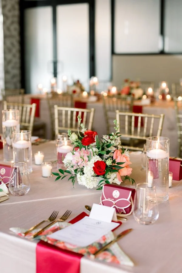 Elegant banquet table decorated with a floral centerpiece of red, pink, and white flowers, surrounded by water glasses with floating candles, pink-themed favor boxes, and place settings with gold flatware and a menu, in a well-lit room with large win