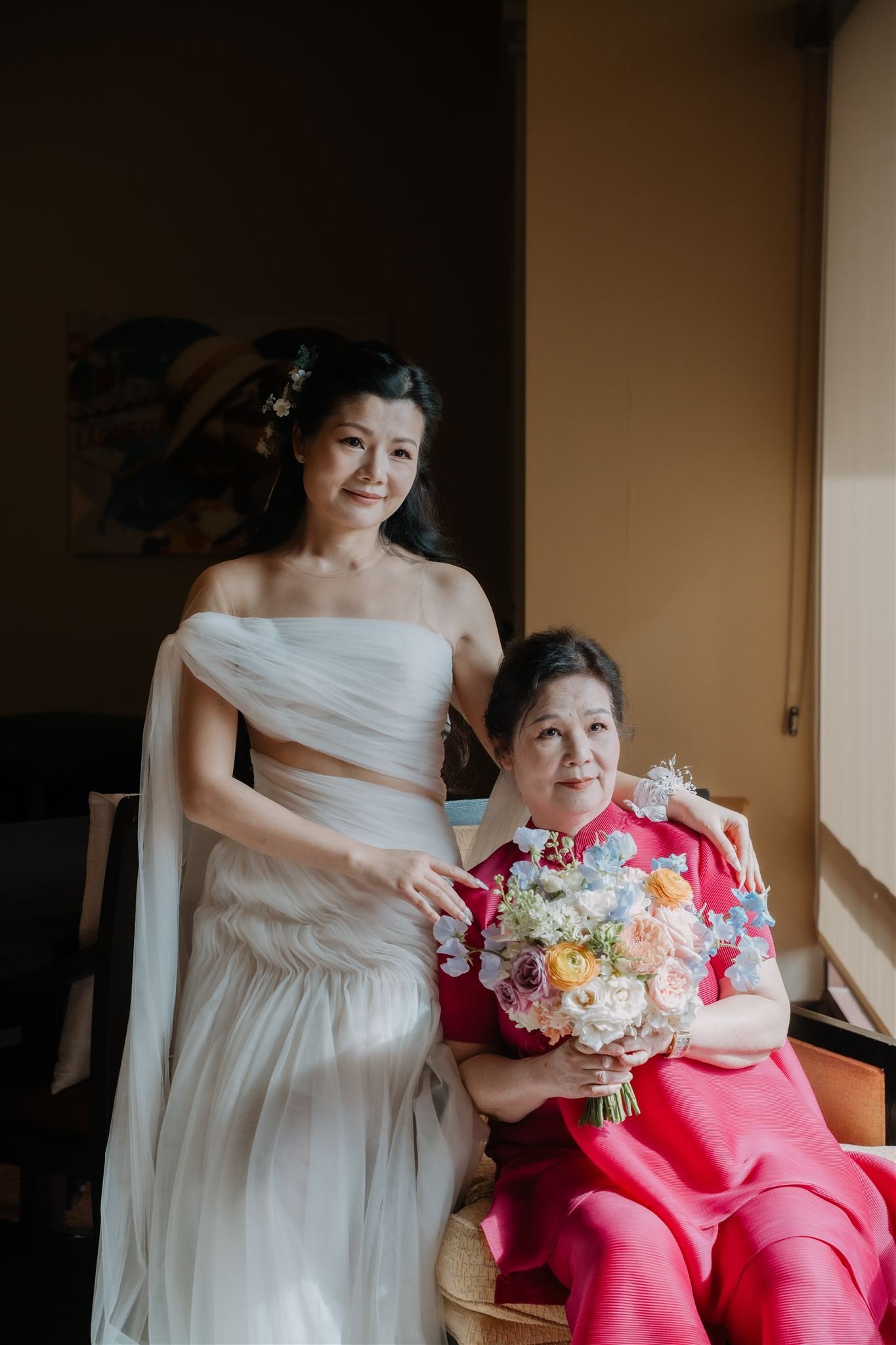 A woman in a white dress is standing next to an older woman sitting on a chair, holding a bouquet of flowers. The woman in the white dress has long dark hair decorated with flowers and is smiling slightly. The older woman is wearing a bright pink out