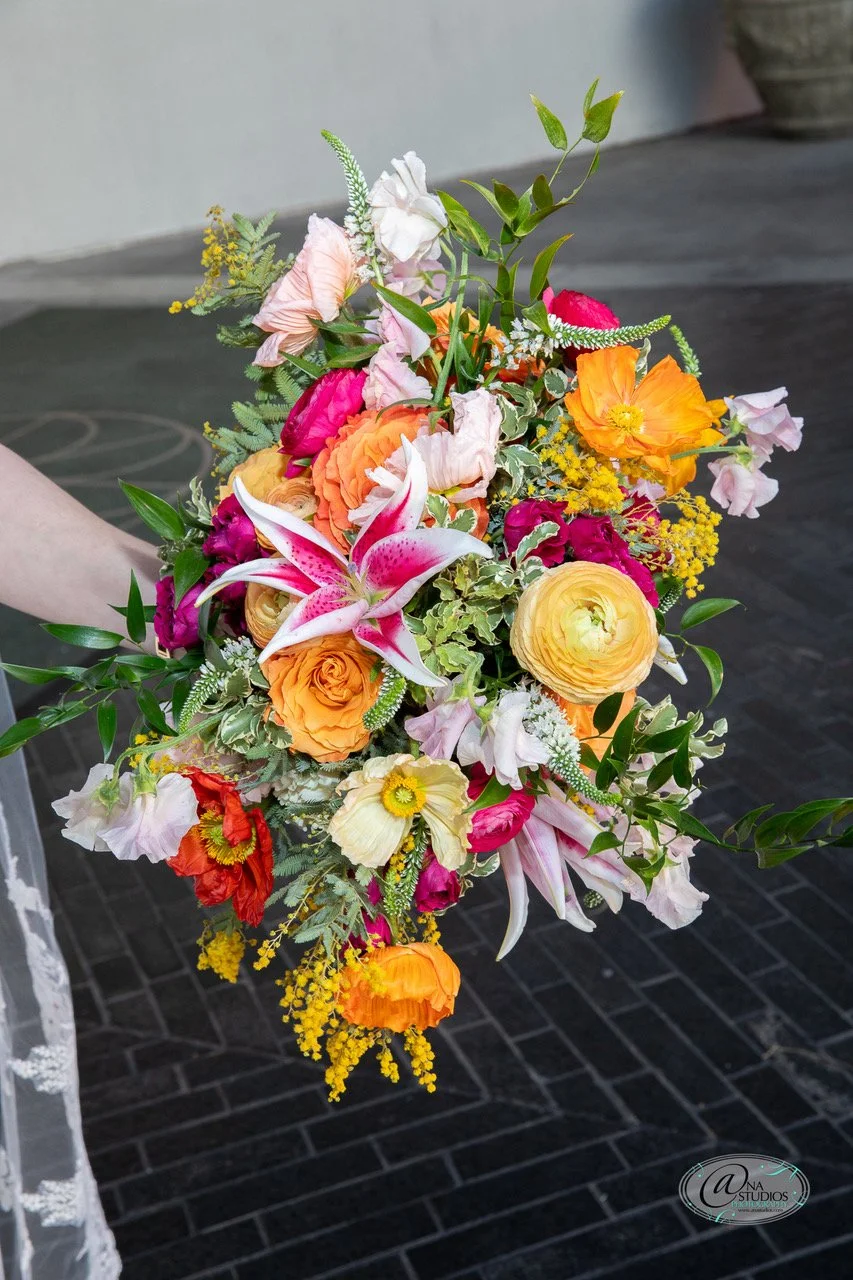 Colorful mixed flower bouquet with pink lilies, orange roses, yellow poppies, and green foliage.