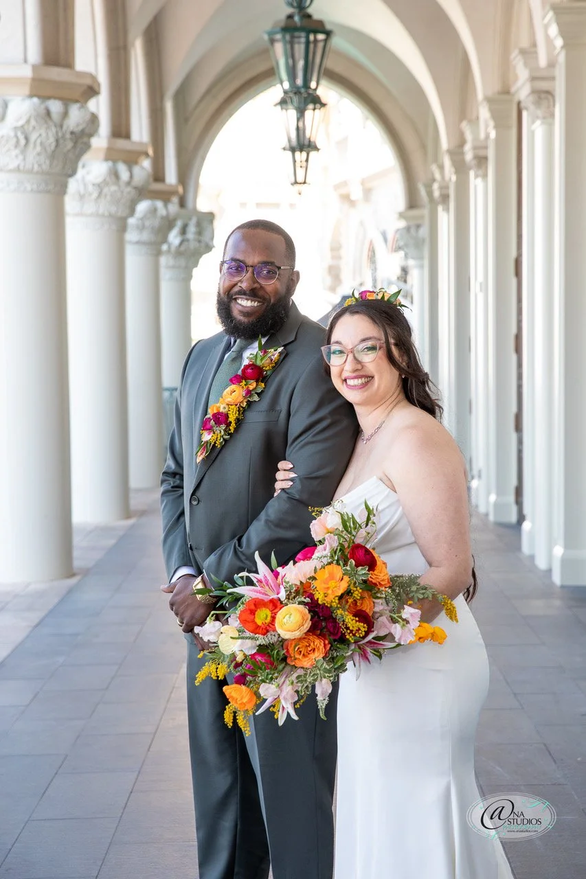 A newlywed couple smiling and posing in a portico with white columns and arches, the man wearing a gray suit and the woman in a white wedding dress holding a colorful bouquet.