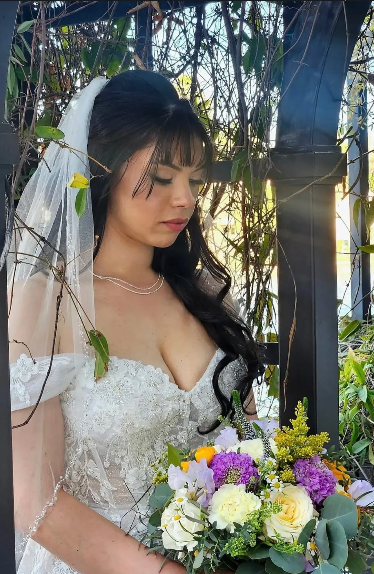 A woman in a white wedding dress holding a bouquet of colorful flowers, standing among vines and greenery with sunlight shining through.
