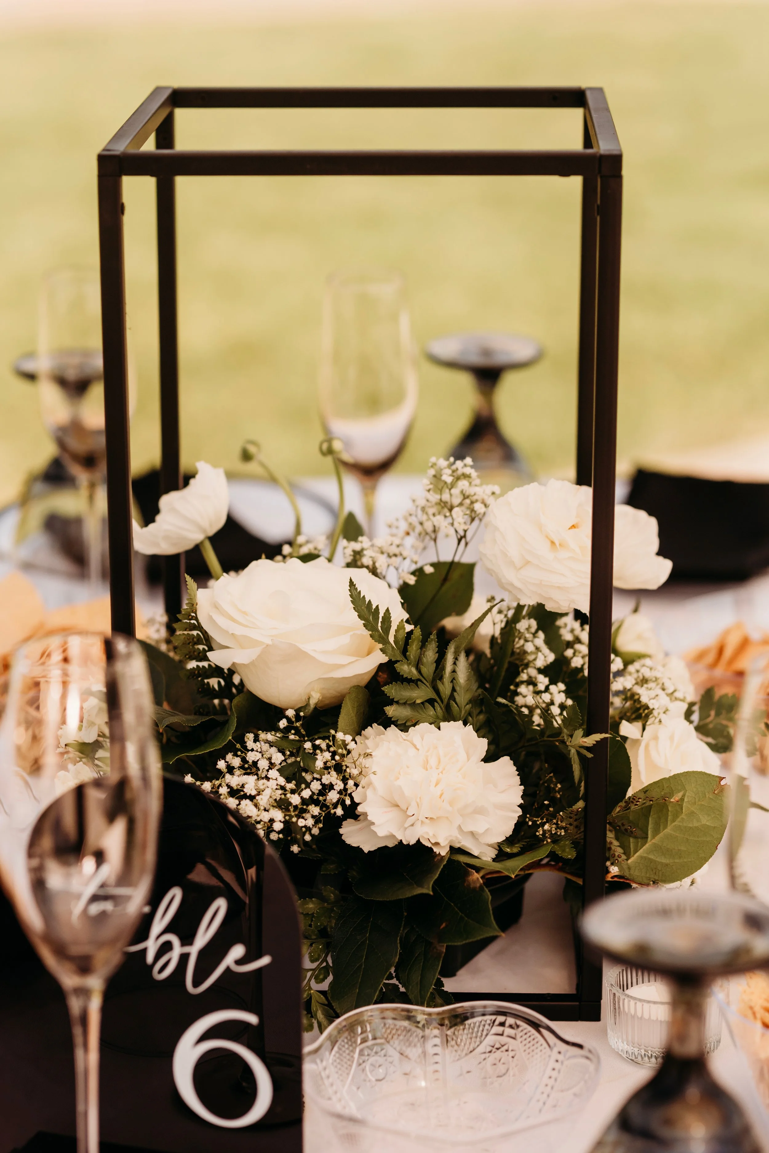 Elegant table centerpiece with white roses, carnations, baby's breath, and greenery inside a black metal frame, surrounded by glassware and table settings at an outdoor event.