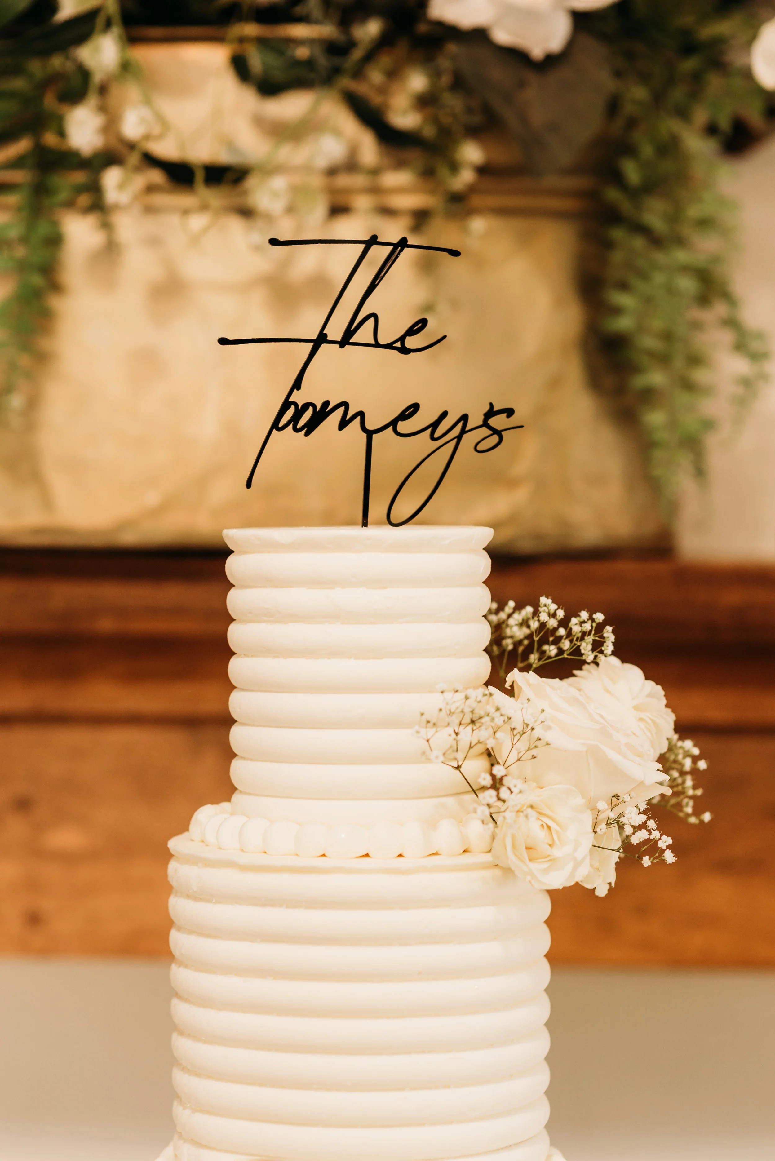 A white wedding cake with horizontal ribbed tiers, decorated with white roses and baby's breath flowers on the side. A black custom cake topper reads 'The Pameys' in elegant script. In the background, there are green foliage and white flowers.