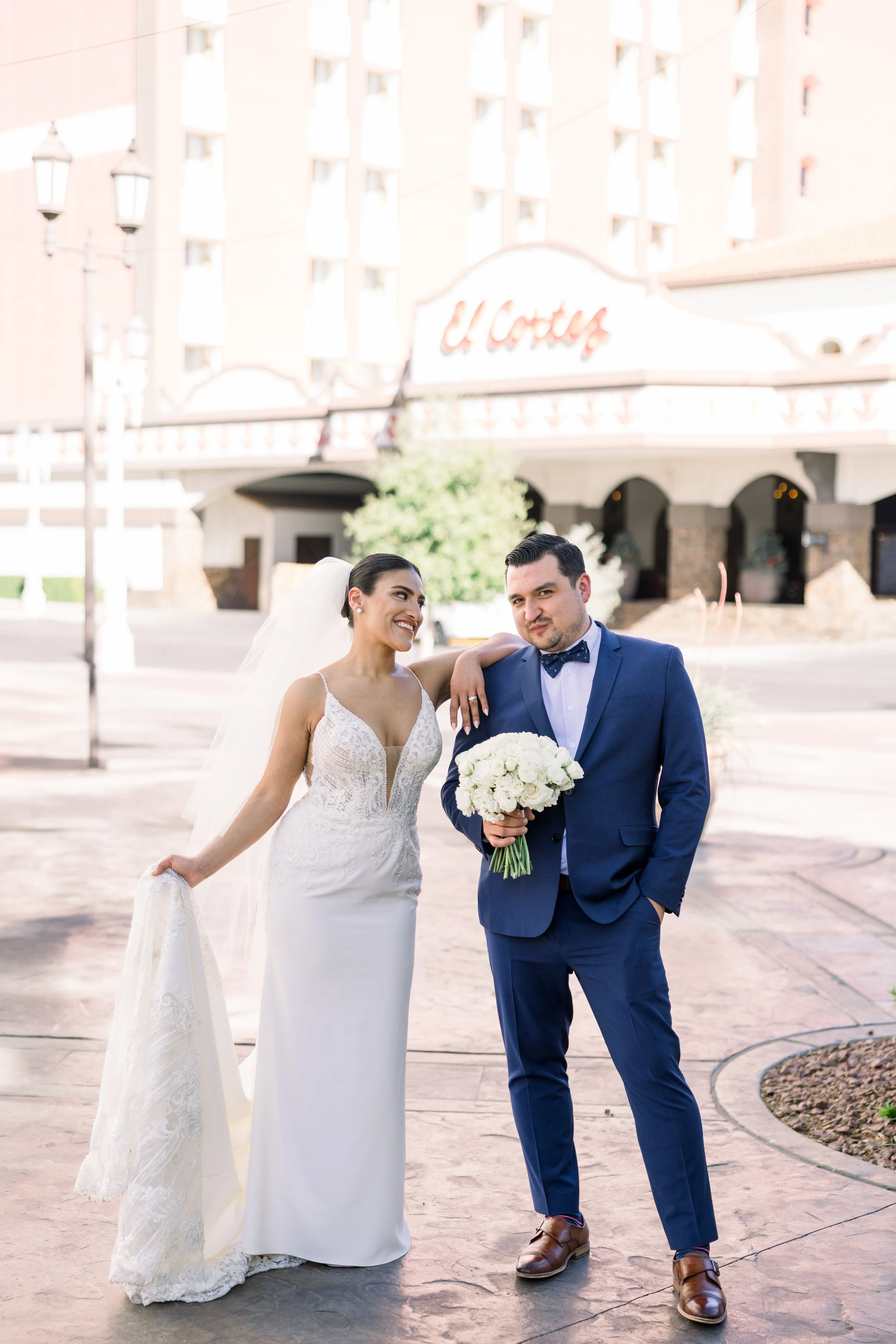 A bride and groom posing under a sign that reads 'U. Corten' outside a building. The bride is holding her dress with her left hand and smiling at the groom. The groom is holding a bouquet of white roses, with his right hand in his pocket, and wearing
