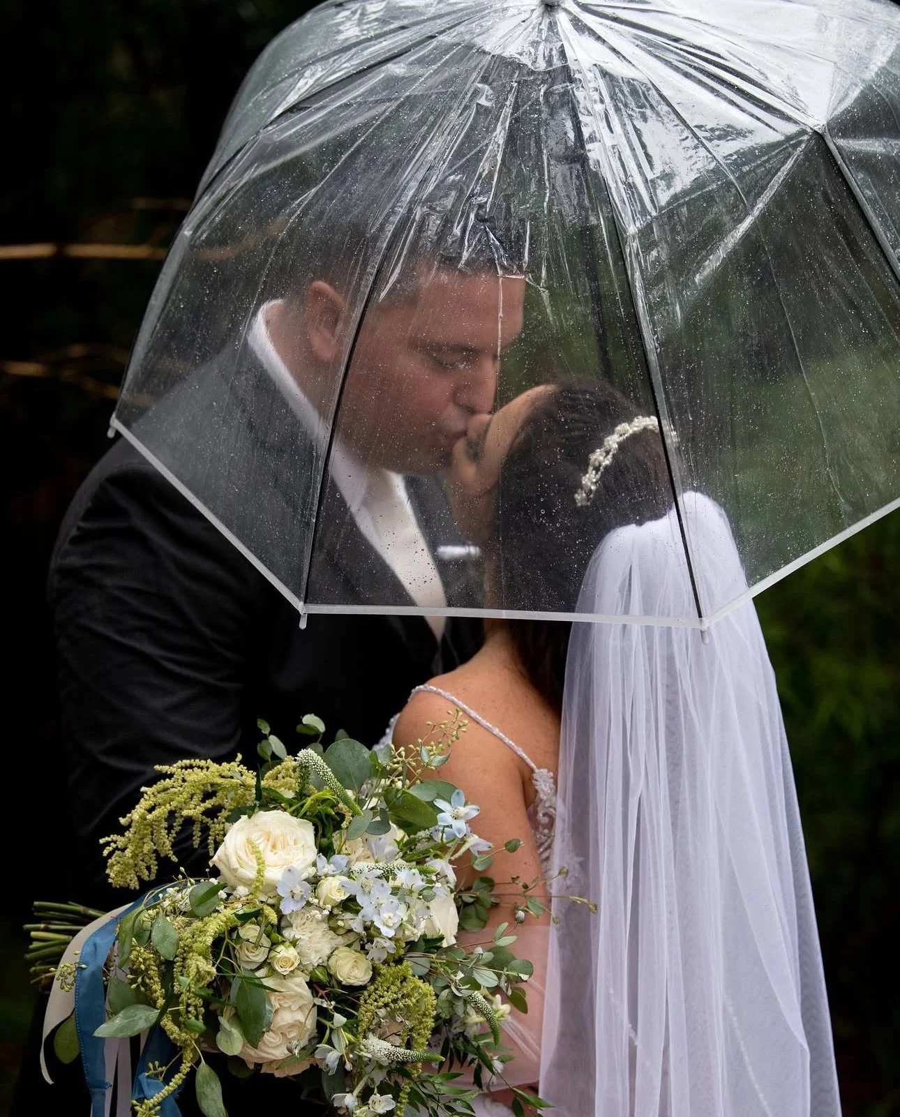 Wedding couple sharing a kiss under a clear umbrella, with the bride holding a large bouquet of white flowers and greenery.