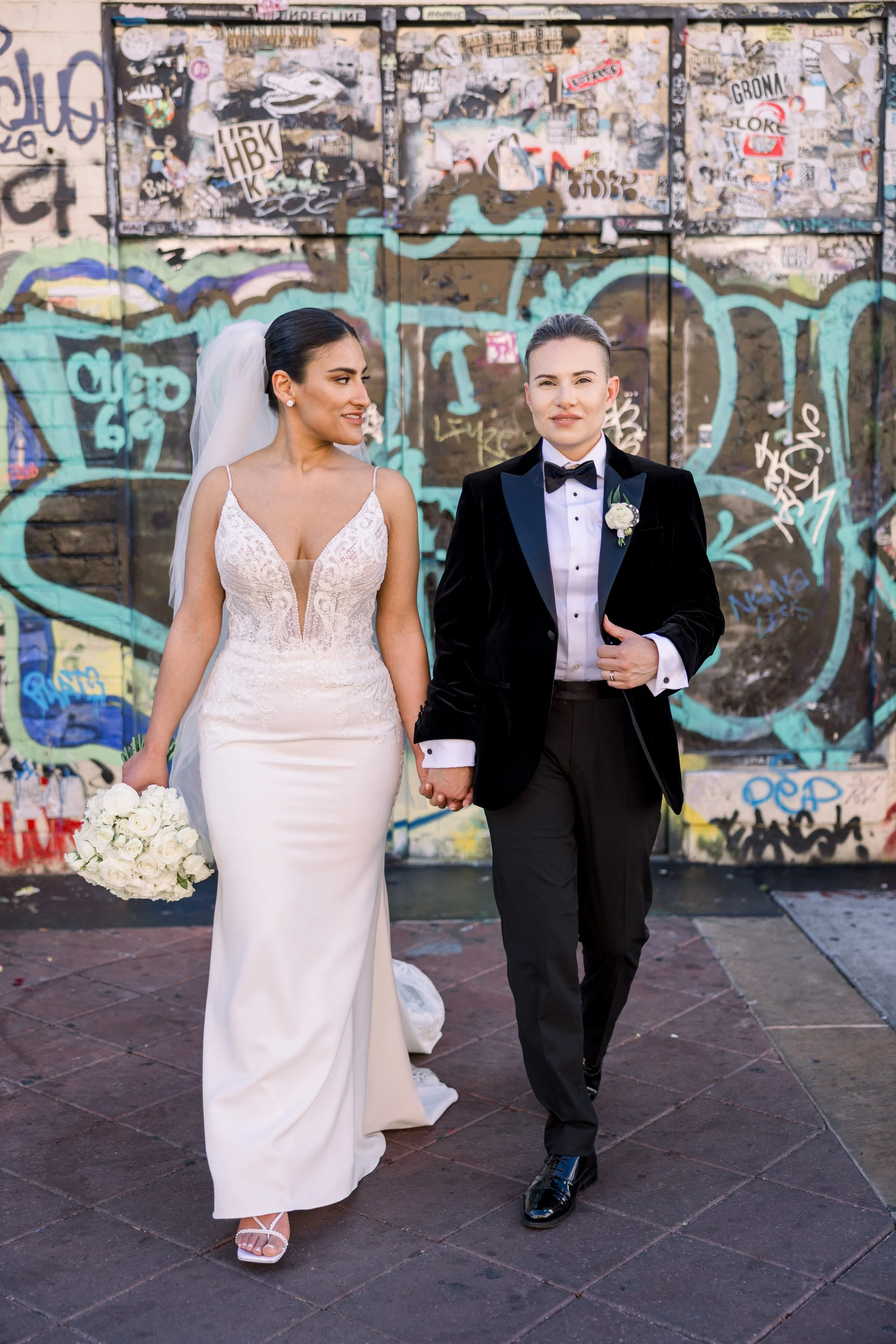 A bride and groom holding hands and walking together in front of a graffiti-covered wall. The bride is wearing a lace wedding dress and veil, holding a bouquet of white roses. The groom is in a tuxedo with a boutonniere, looking at the bride.