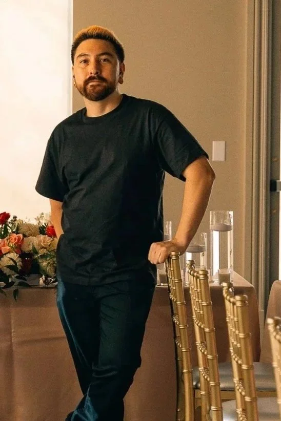 A man with dark hair and a beard standing indoors beside gold-colored chairs and a table with water glasses, with a flower arrangement and a beige wall in the background.