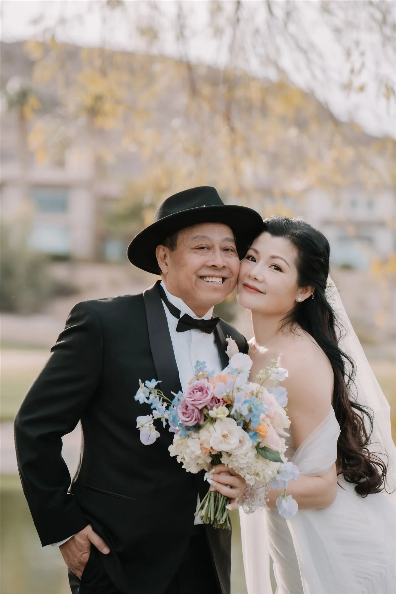 A bride and an older man, possibly her father, smiling and posing together outdoors during daytime. The bride is holding a bouquet of colorful flowers and wears a white wedding dress, while the man is dressed in a tuxedo with a bow tie and a black ha