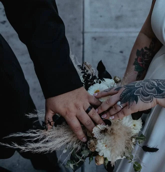 Two people holding hands over a bouquet of flowers, including white blooms and dark feathers, in front of a gray wall.