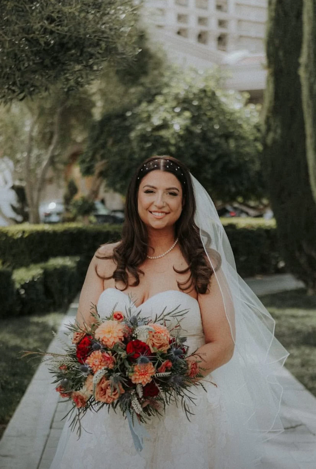 A smiling bride with dark brown hair wearing a strapless white wedding gown, pearl necklace, and veil, holding a bouquet of red and peach flowers, standing outdoors in a park with trees and buildings in the background.