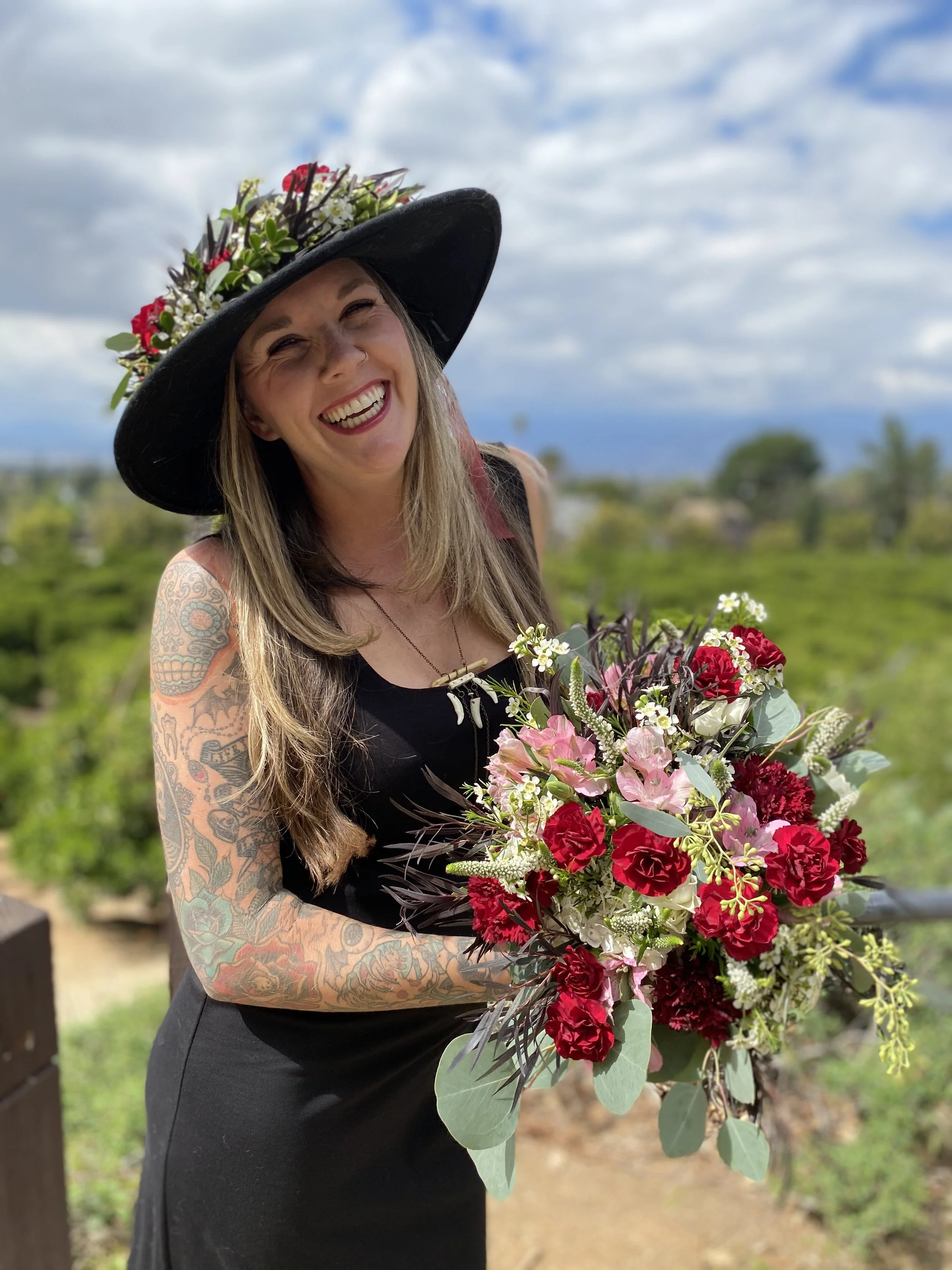 Woman with long hair wearing a large black hat decorated with flowers, smiling and holding a large bouquet of pink, red, and white flowers outdoors, with a scenic background of trees and a partly cloudy sky.