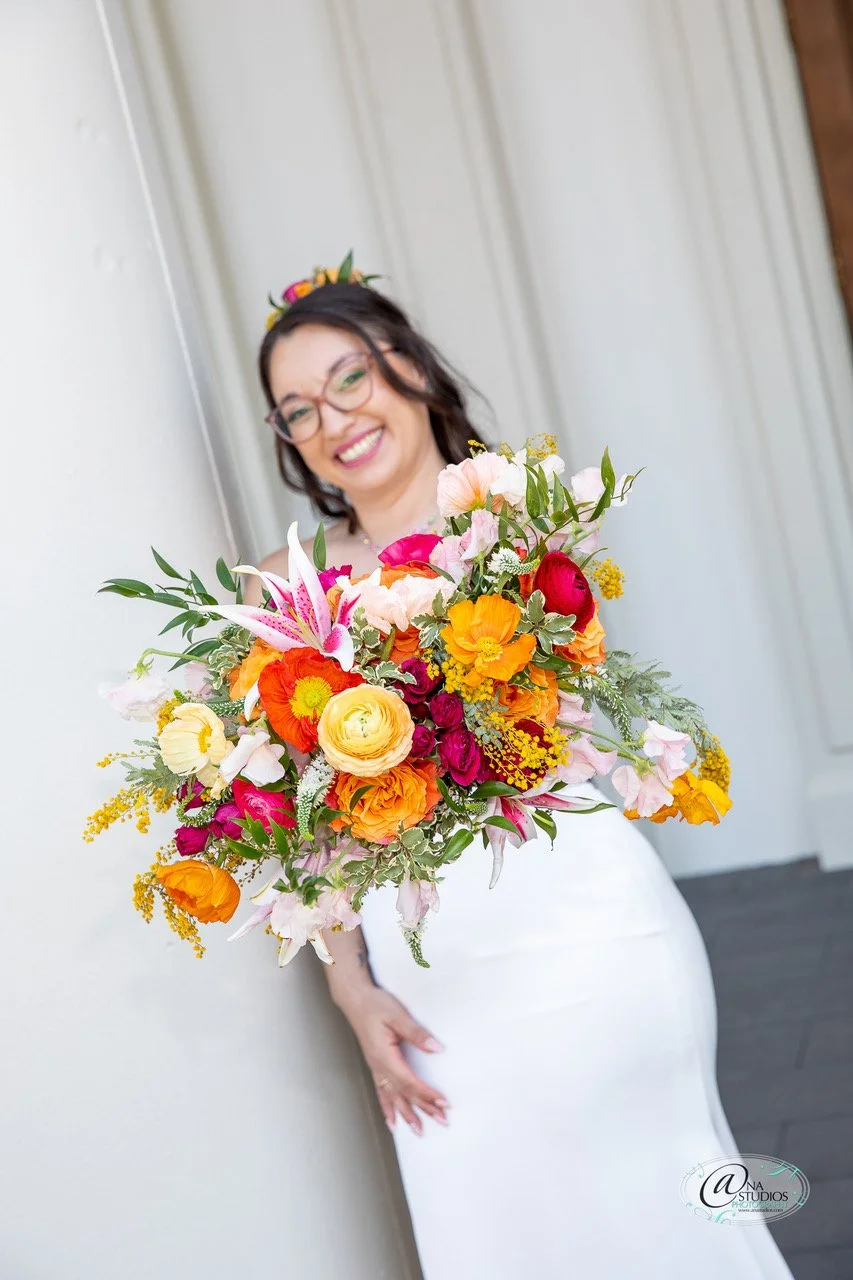 A woman in a white dress holding a large, colorful bouquet of flowers, smiling, with glasses and dark hair, standing indoors against a white wall.