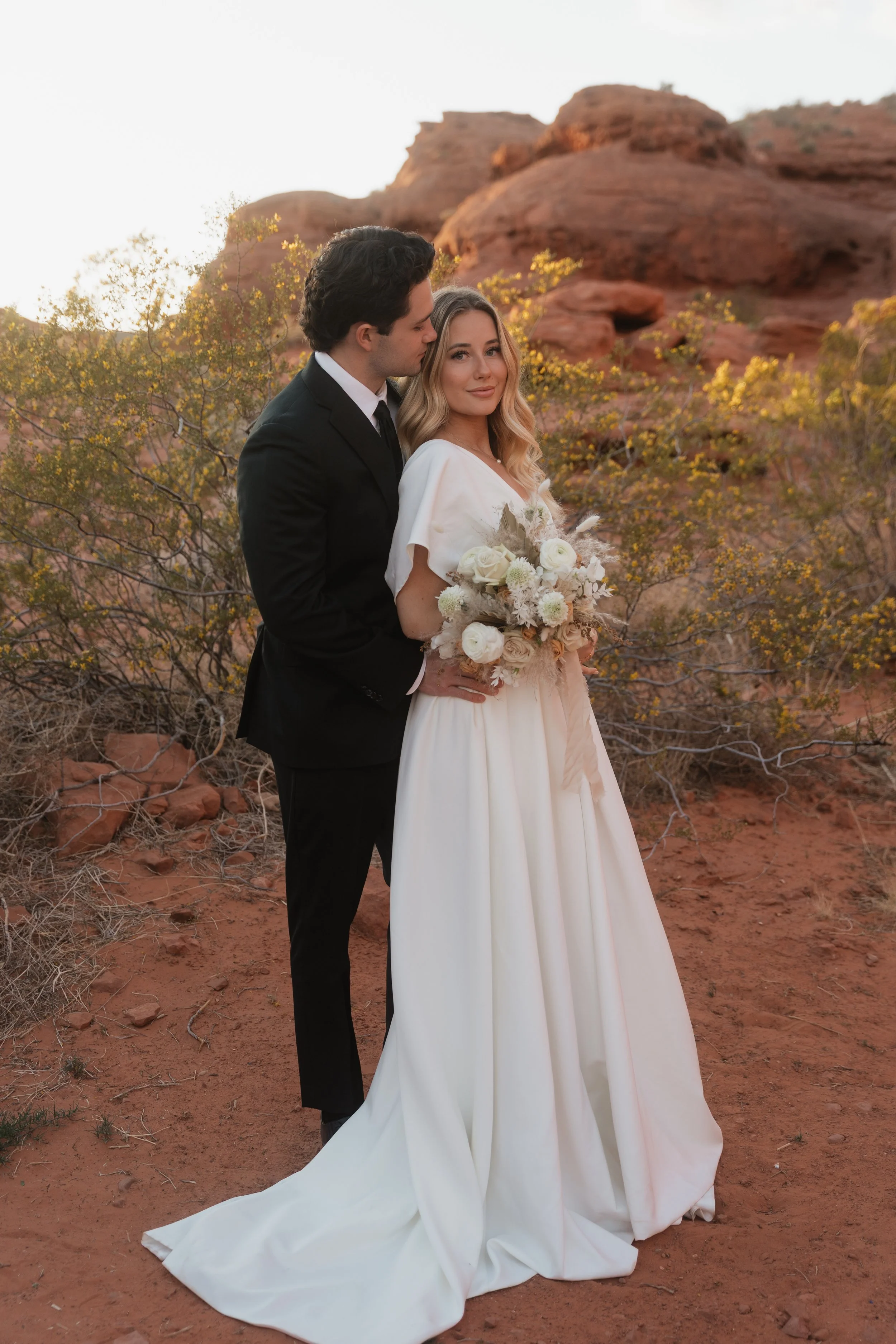 A bride and groom standing close together outdoors in a desert landscape with red rocks and yellow foliage, during sunset. The bride wears a white wedding gown and holds a bouquet, while the groom is in a black suit.