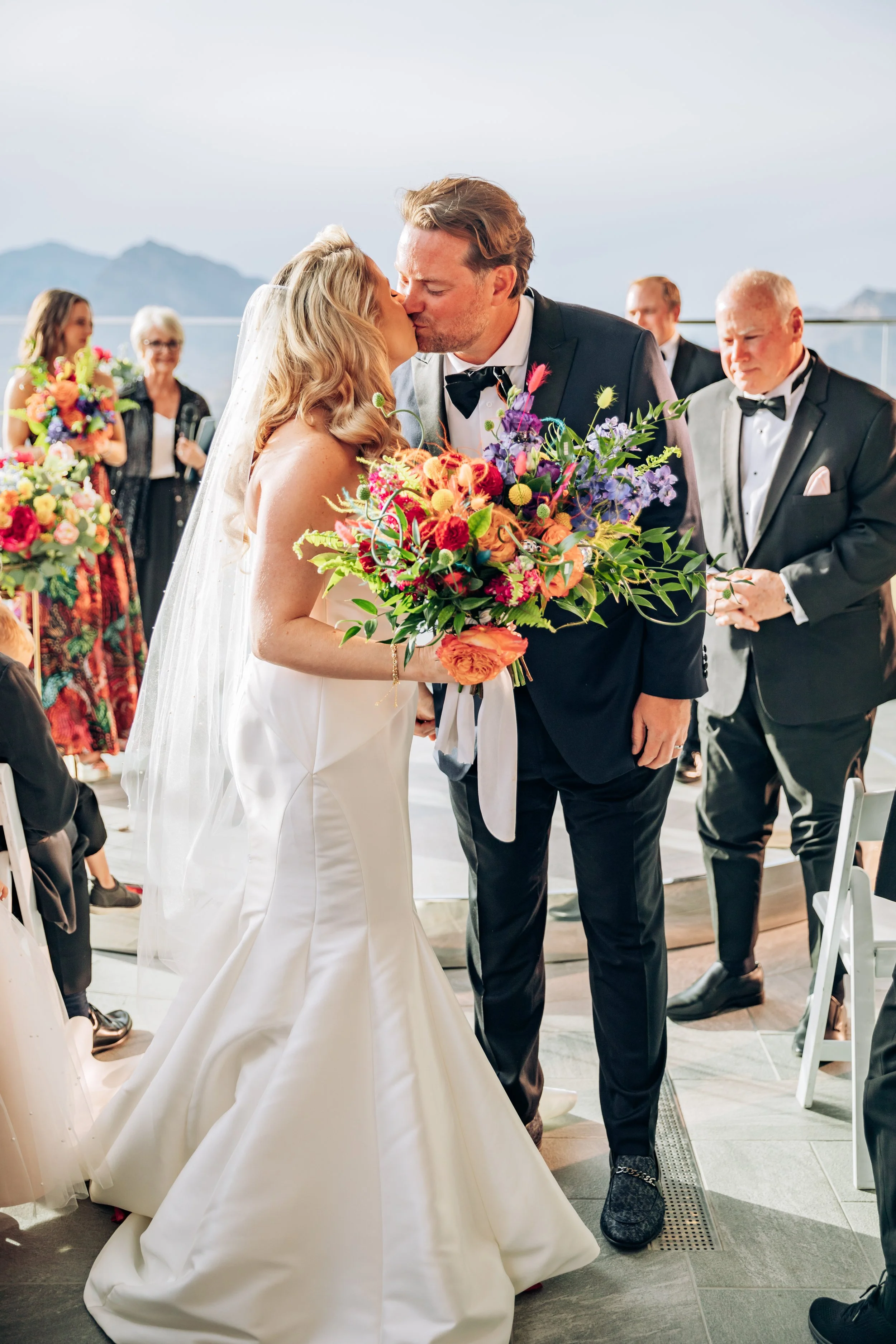 A bride and groom kiss during their wedding ceremony on a scenic oceanfront with mountains in the background. The bride holds a colorful bouquet, and guests are seen in the background celebrating.